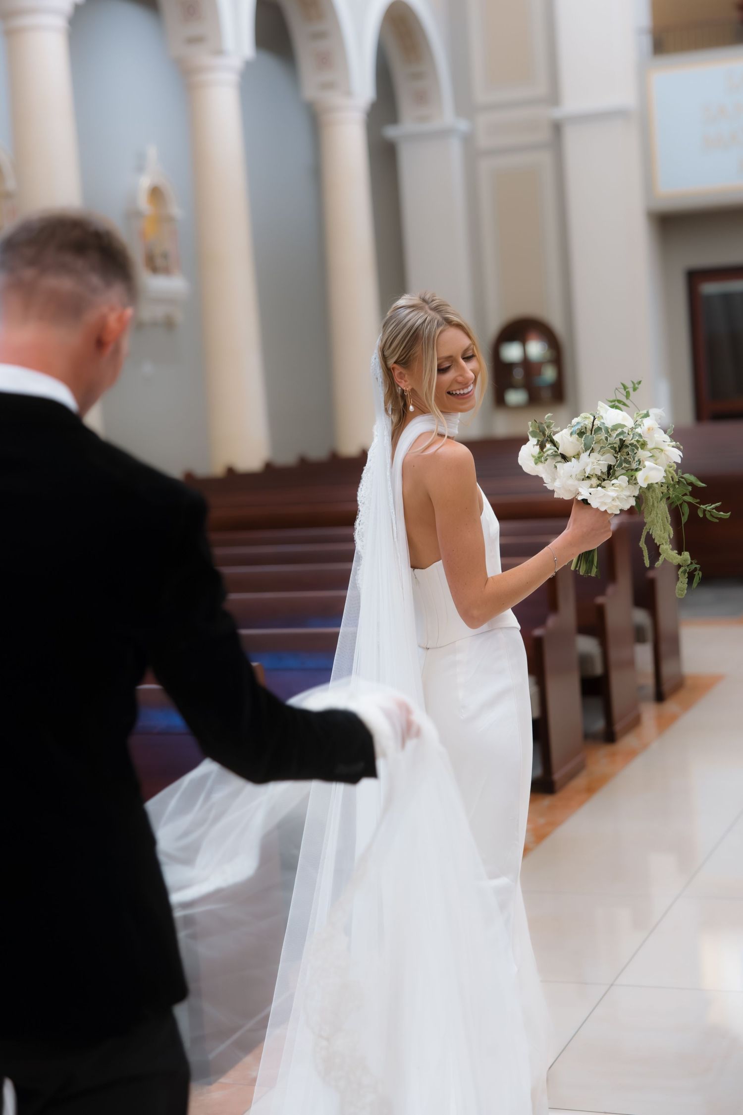 Smiling bride in white halter gown holds bouquet while walking down church aisle with groom.