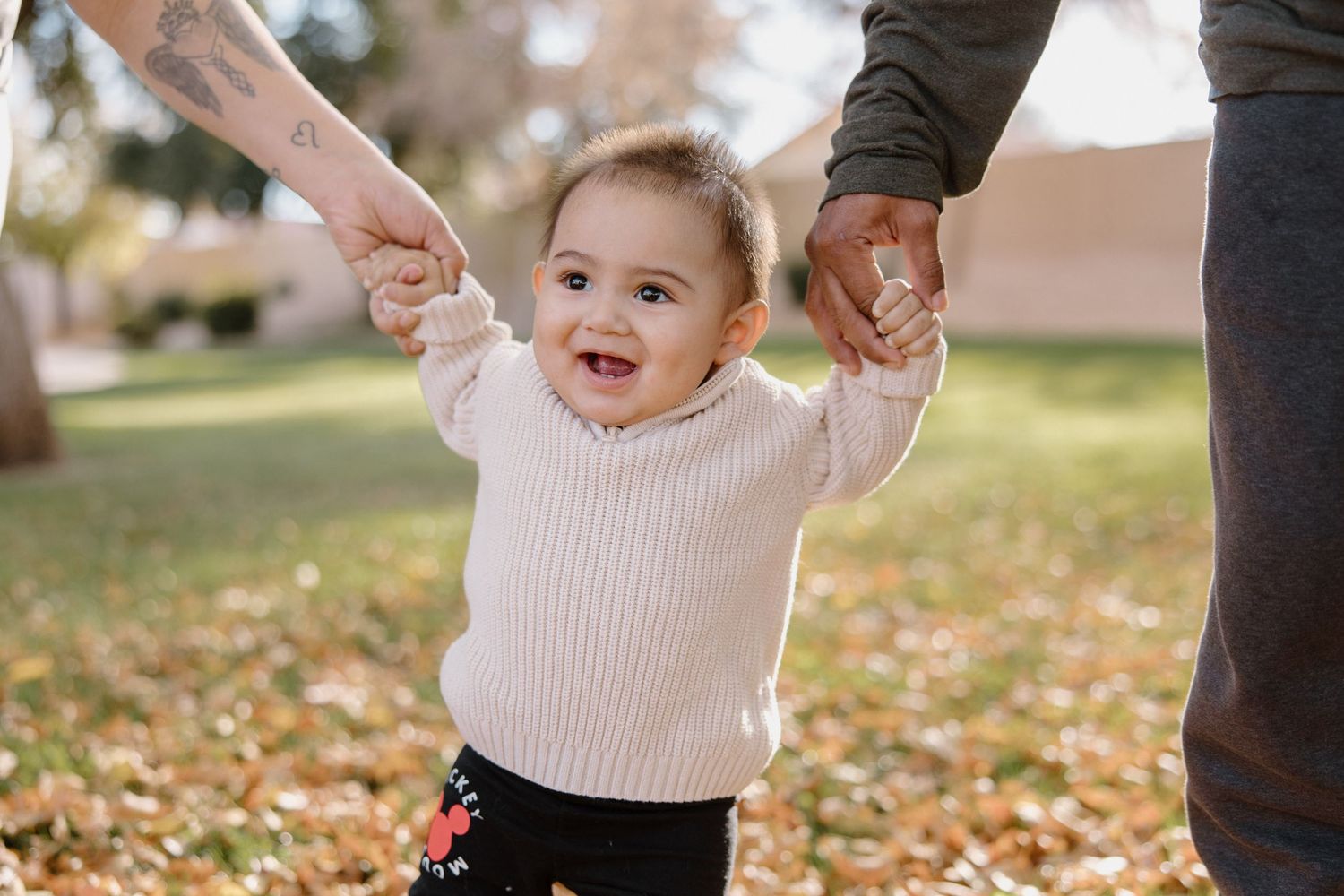 A smiling baby in a cream sweater takes first steps outdoors, held by two adults hands among autumn leaves.