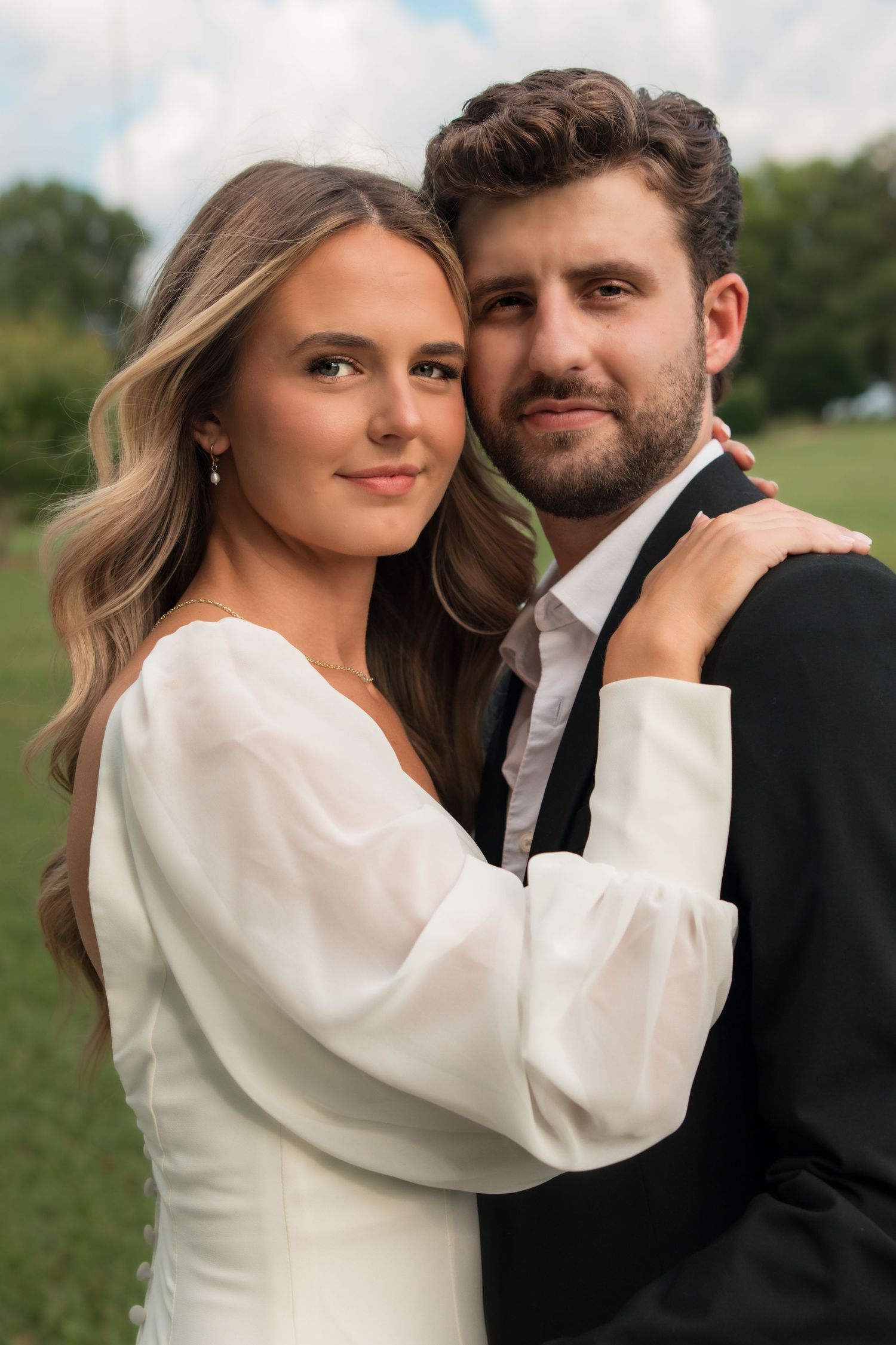 A couple in wedding attire embraces outdoors, the bride in white and groom in black suit, smiling at the camera.