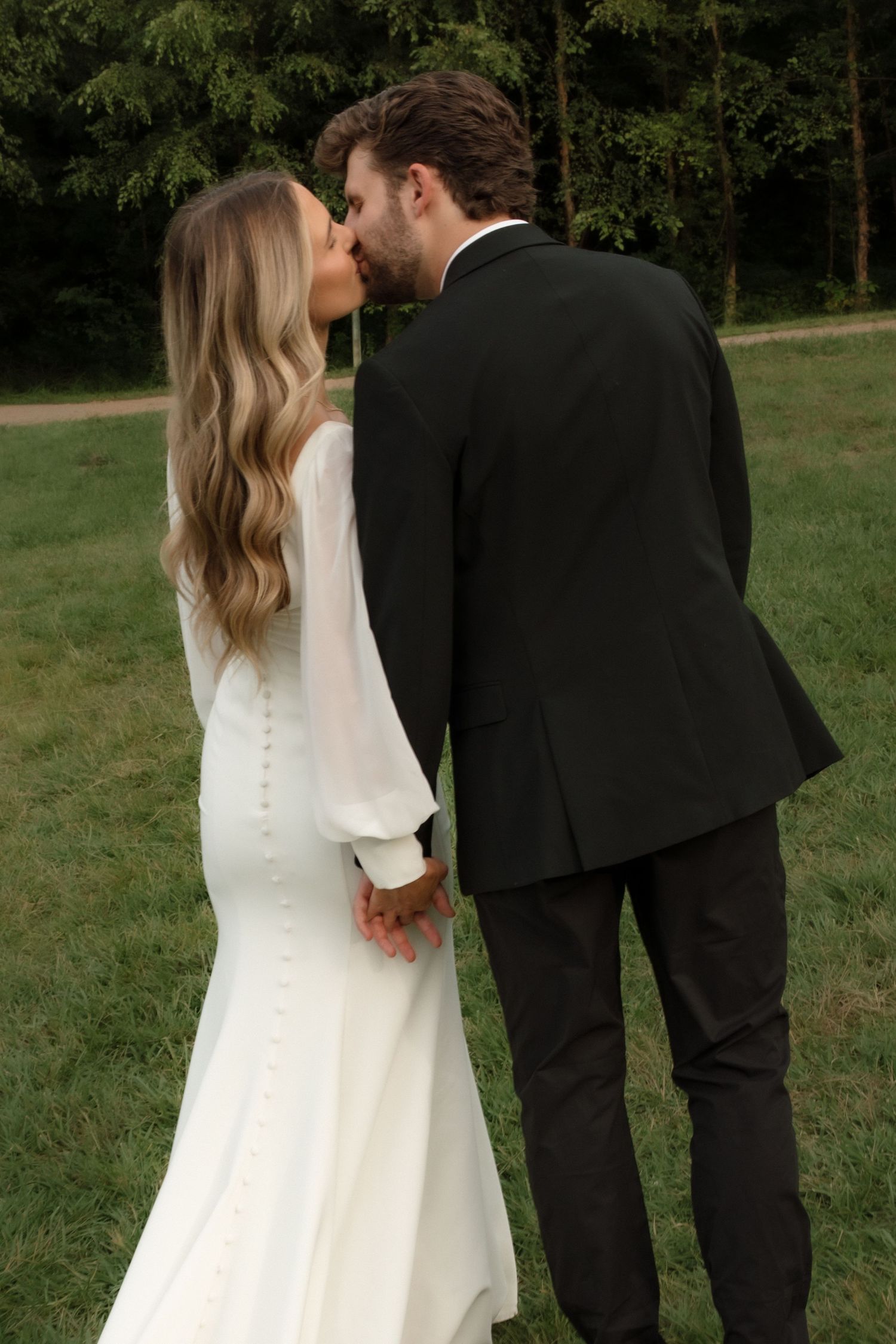 Bride and groom share a tender kiss from behind, bride in button-back white gown, groom in black tuxedo.