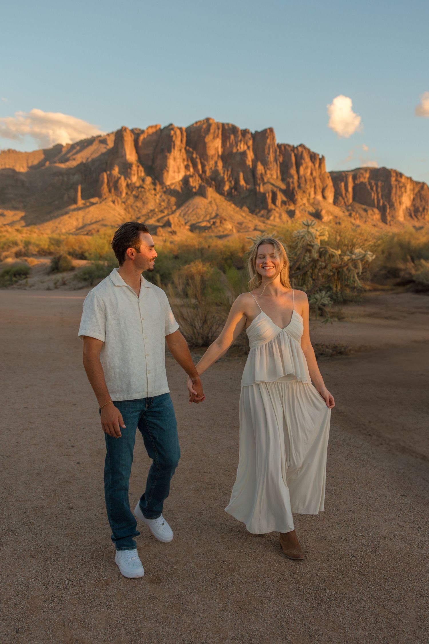 Couple holding hands walking in desert landscape with Superstition Mountains glowing at golden hour sunset.