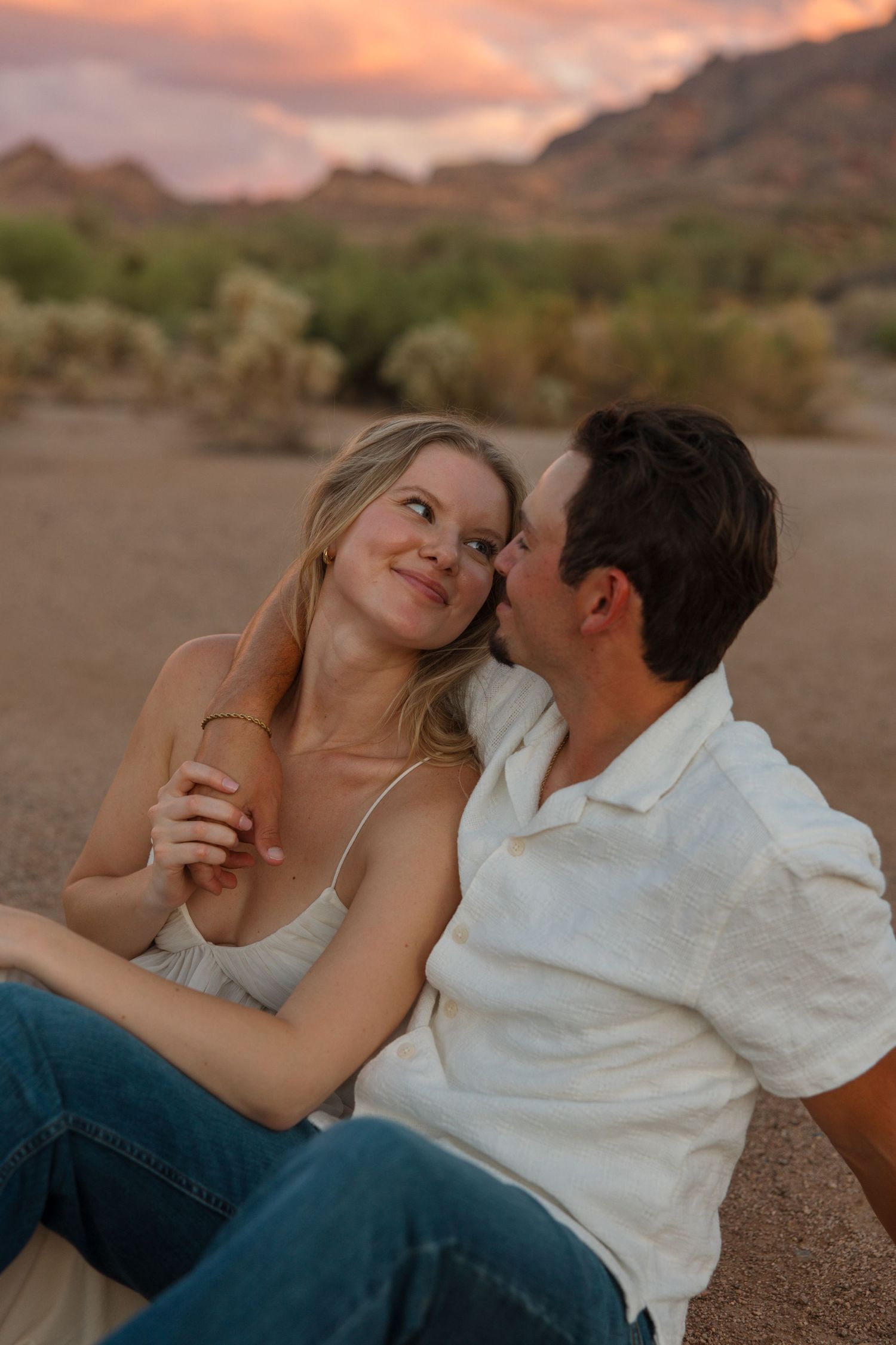 Couple sitting together in desert landscape at sunset, woman smiling up at man as he leans in close, mountains in background.