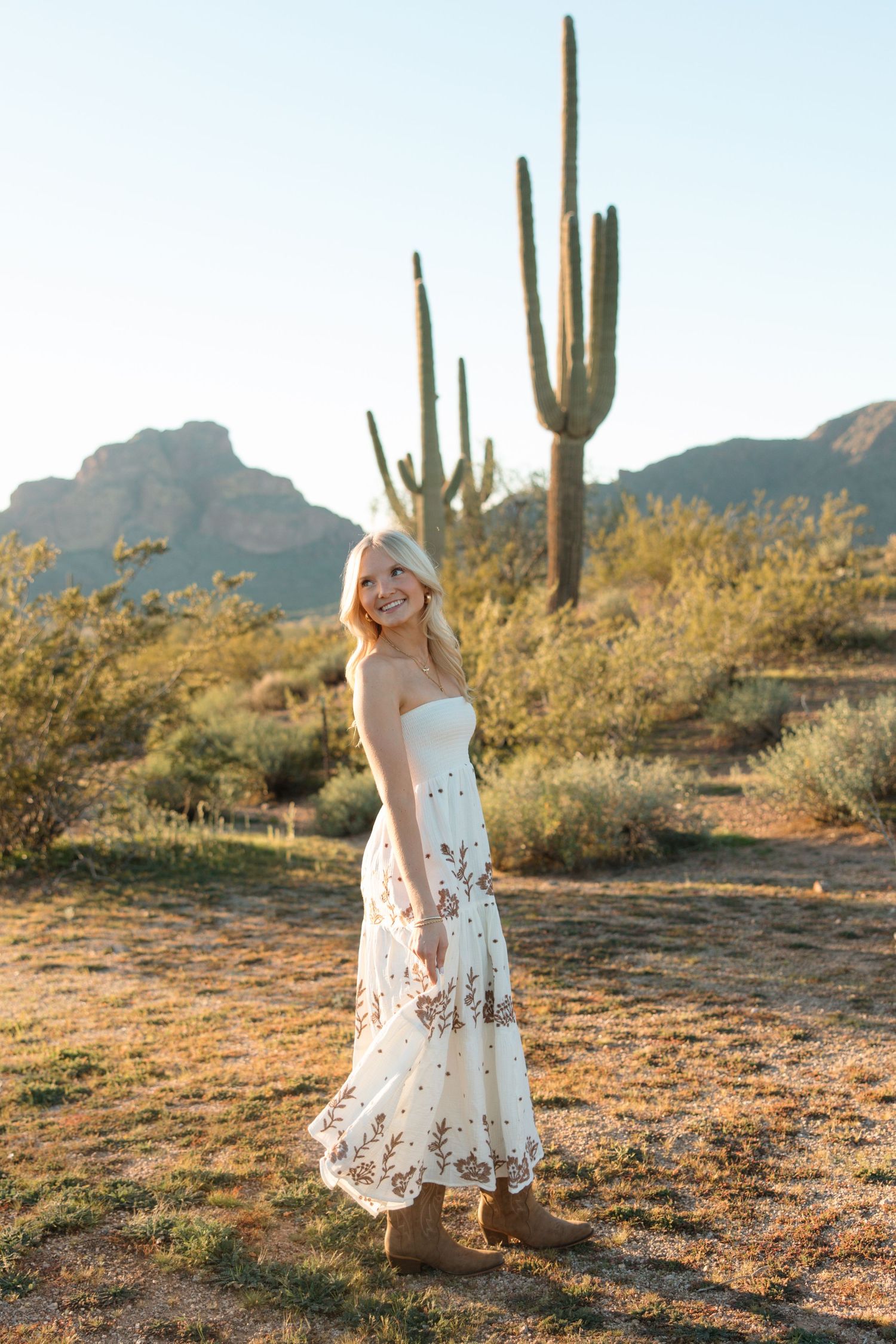 Smiling blonde woman in white floral maxi dress and cowboy boots posing in Arizona desert with saguaro cactus.