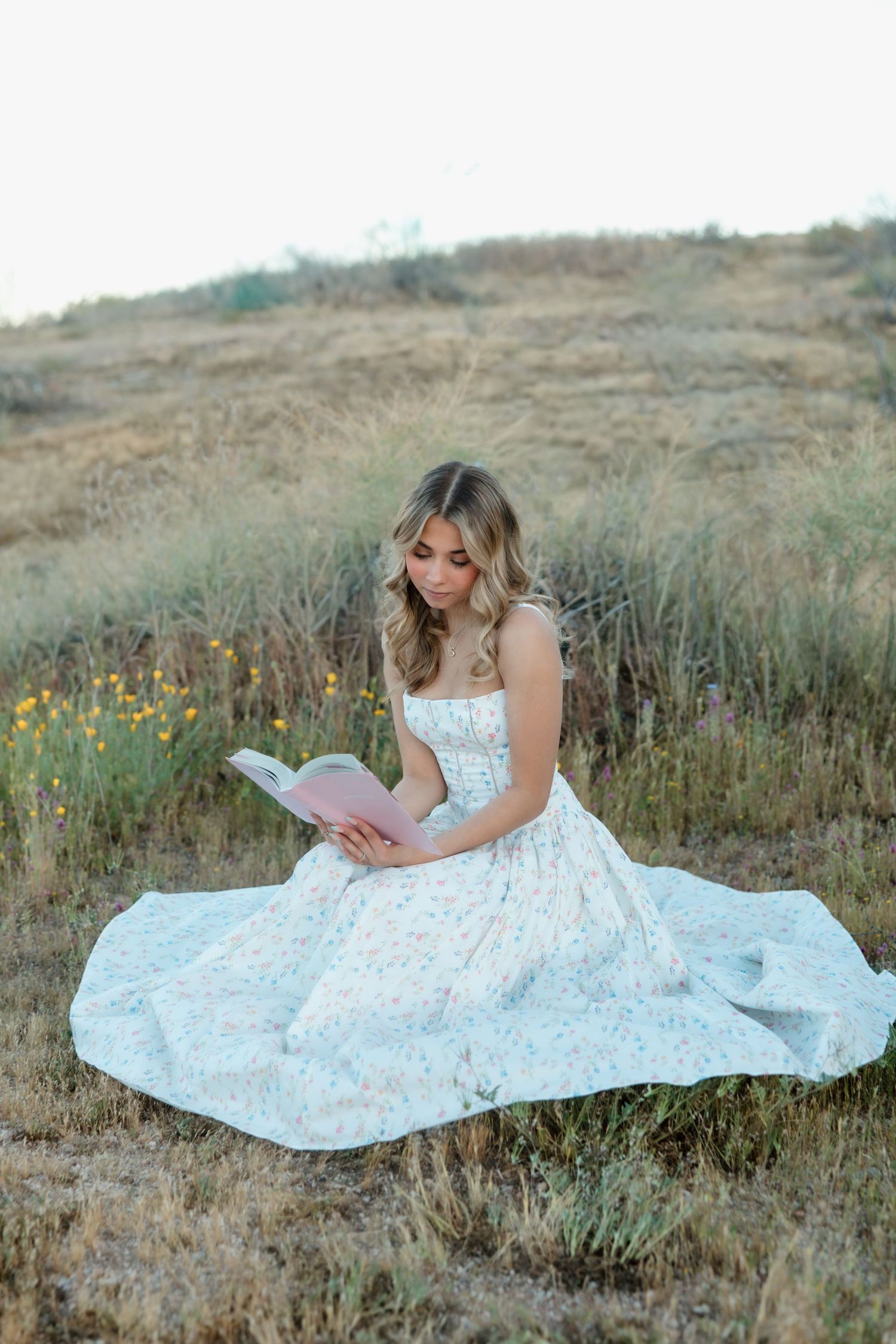 Young woman in floral sundress sitting in a field reading a pink book surrounded by wildflowers.