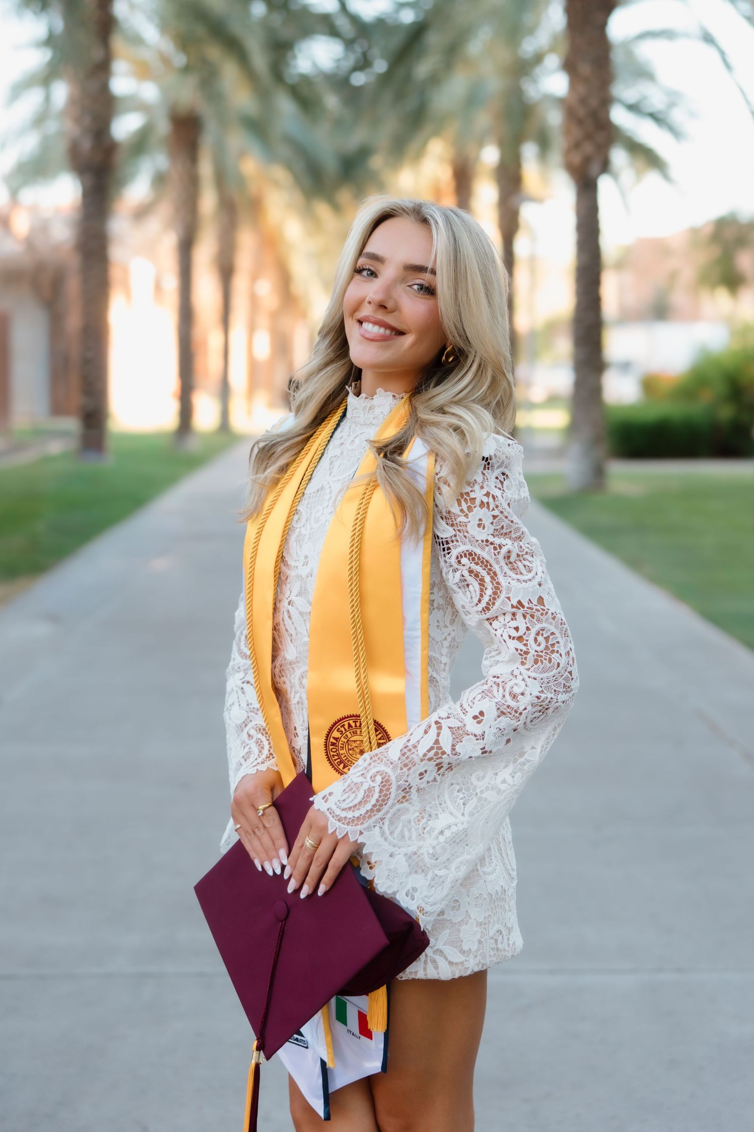 Blonde graduate smiles holding maroon cap wearing white lace dress and gold honor stole among palm trees.