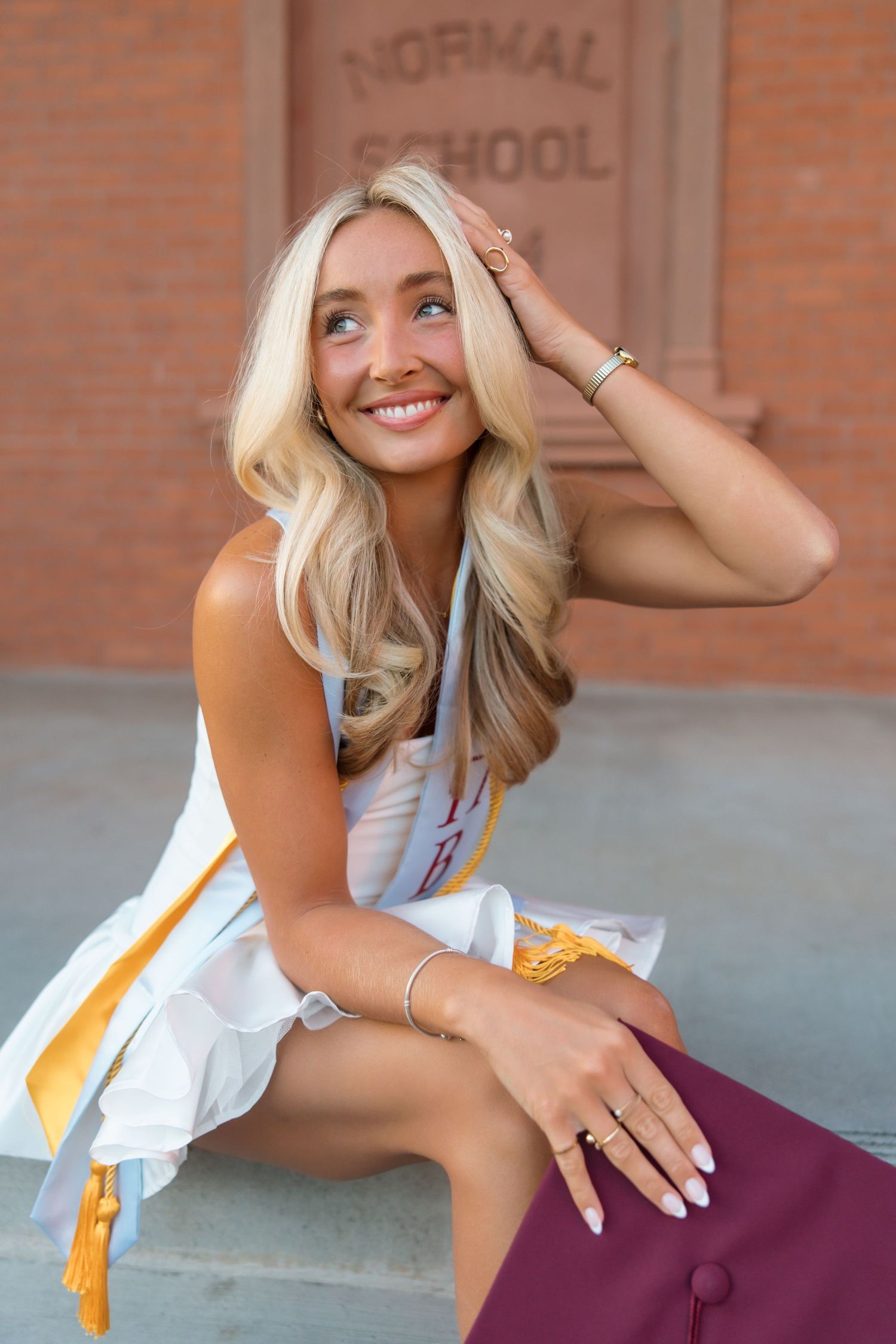 Smiling blonde graduate in white dress and gold honor sash sits outside Normal School building with maroon cap.