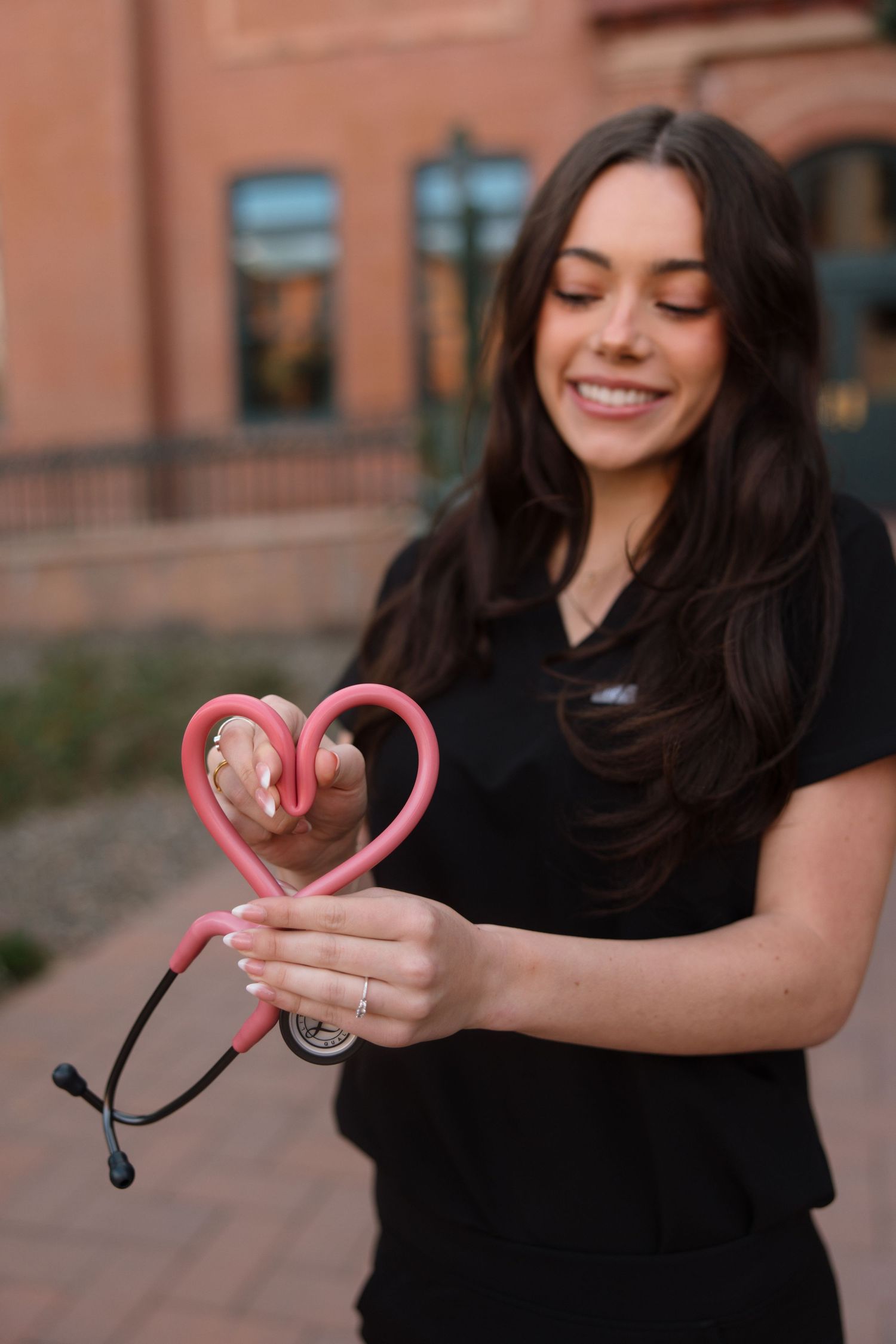 Nursing graduate in black scrubs forming heart shape with pink stethoscope outdoors on campus.