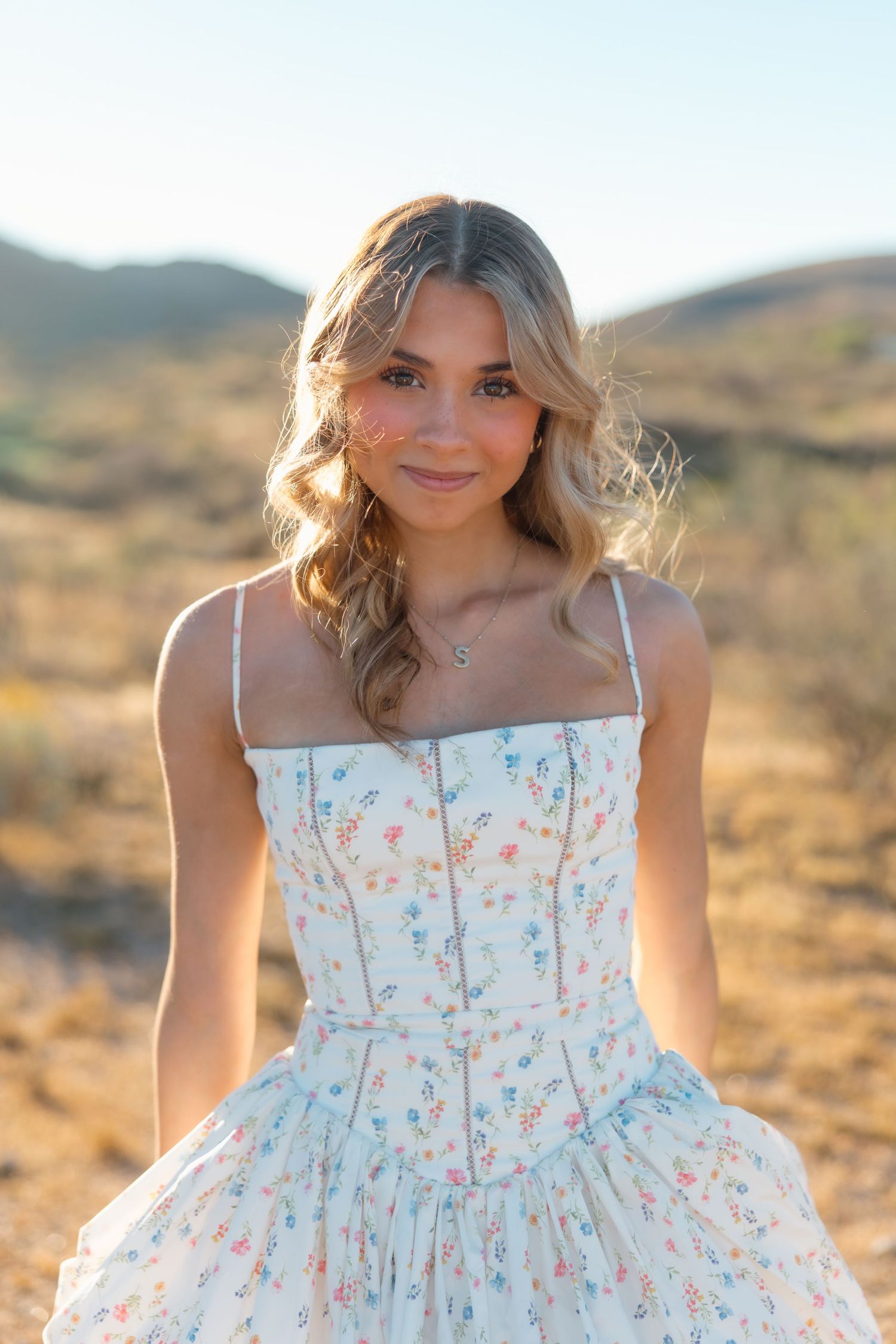 Smiling blonde woman in white floral corset dress sitting in desert landscape.
