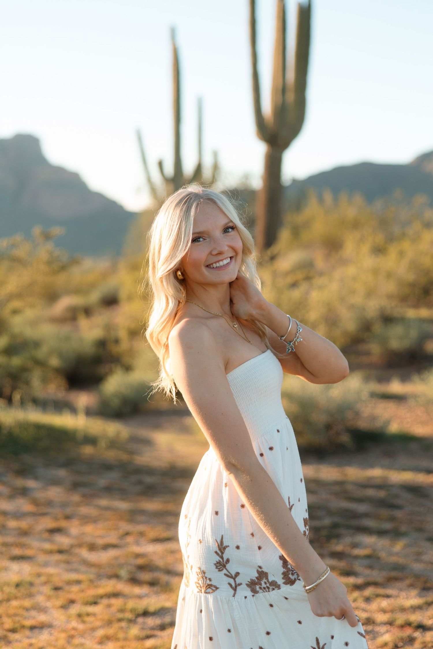 Blonde woman in white floral strapless dress smiling in Arizona desert with saguaro cactus.