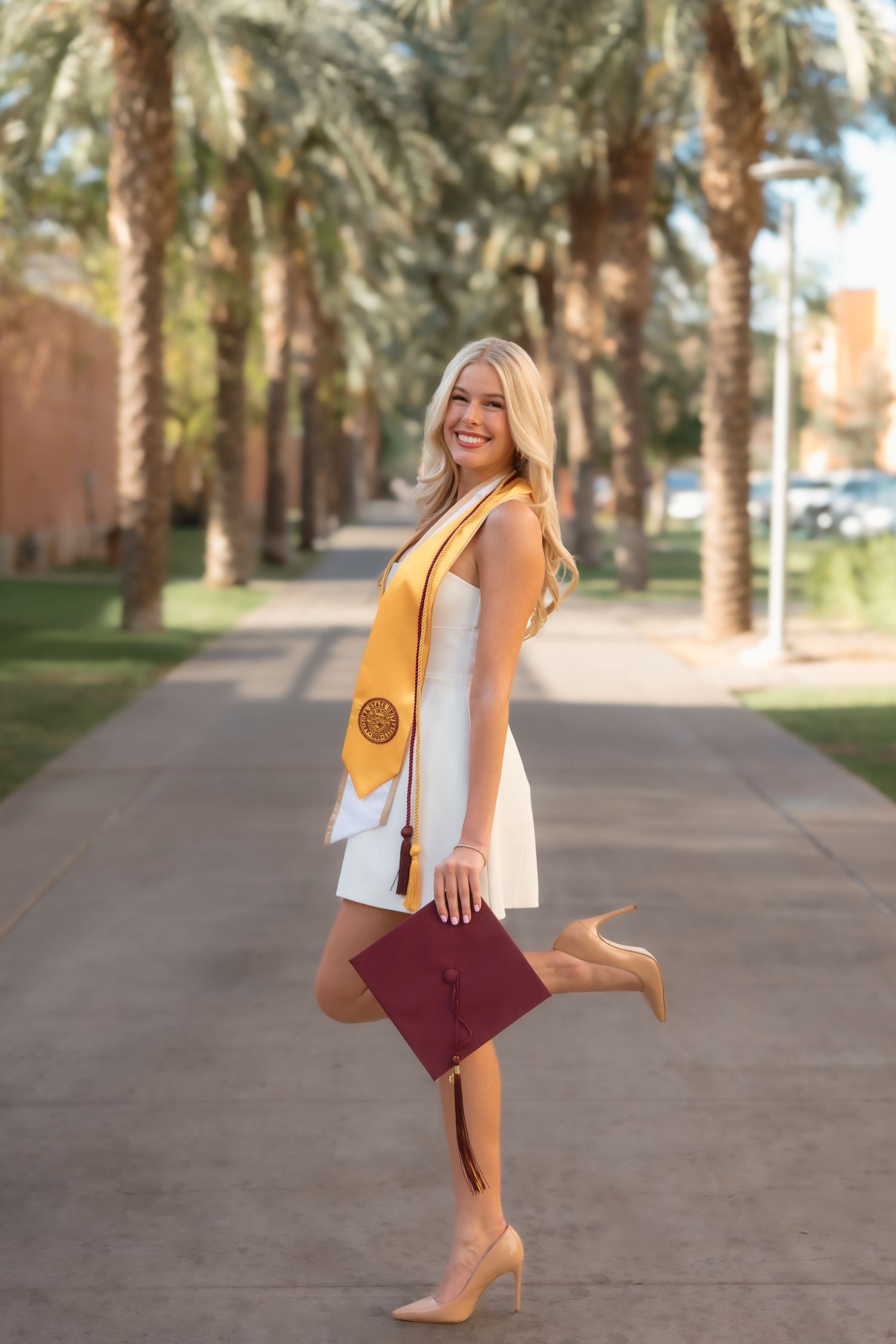 Blonde graduate poses playfully in white dress with gold ASU honor stole holding maroon cap outdoors.