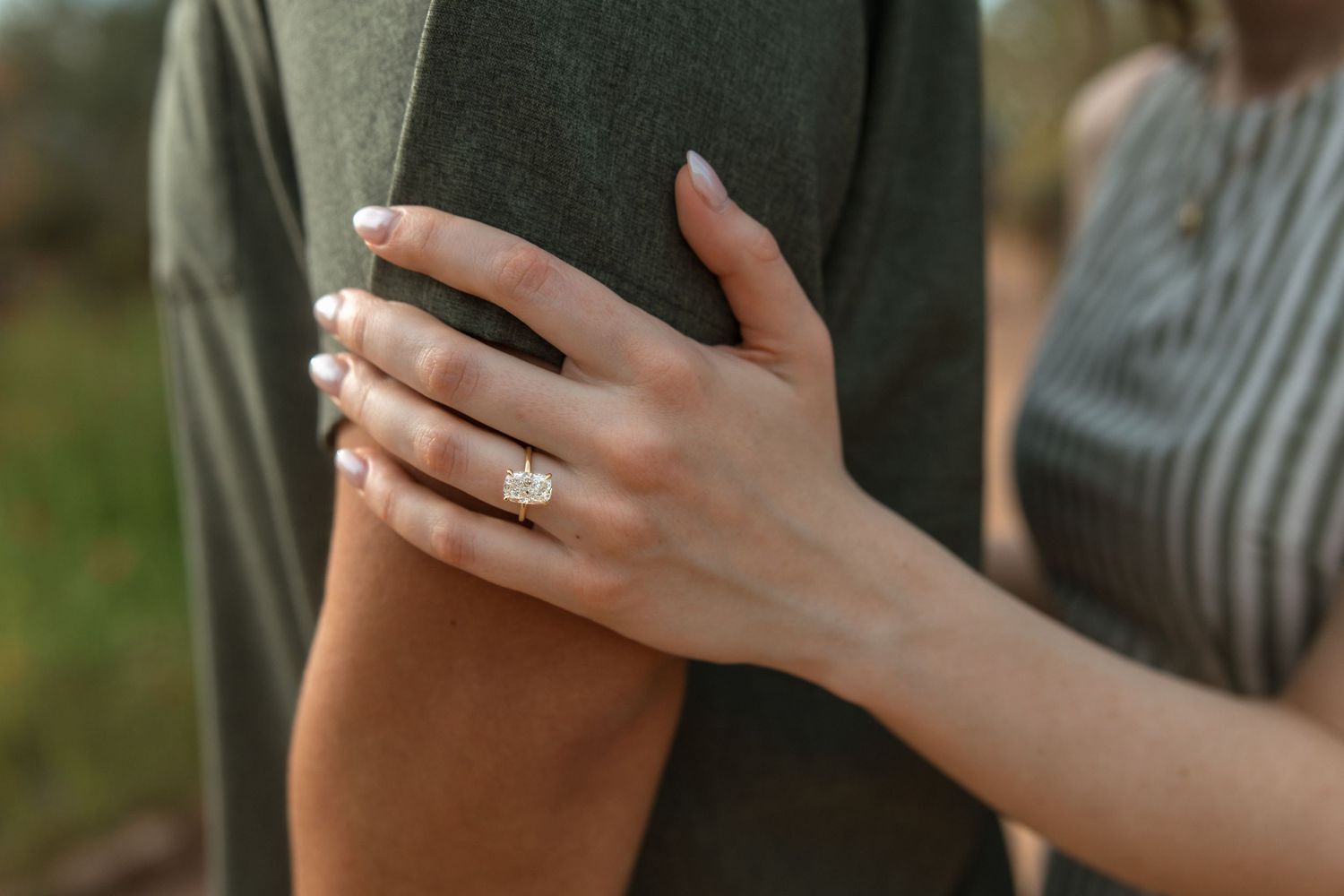 Close-up of a womans hand with a elegant diamond engagement ring resting on a mans green shirt sleeve.