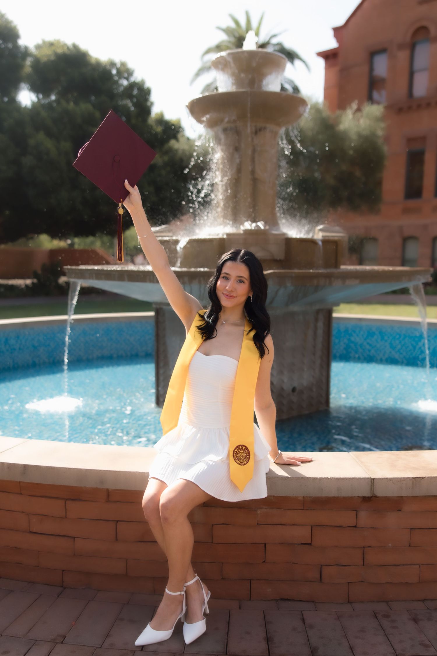 Young woman in white dress and yellow graduation stole raises diploma by a fountain, celebrating her graduation day.