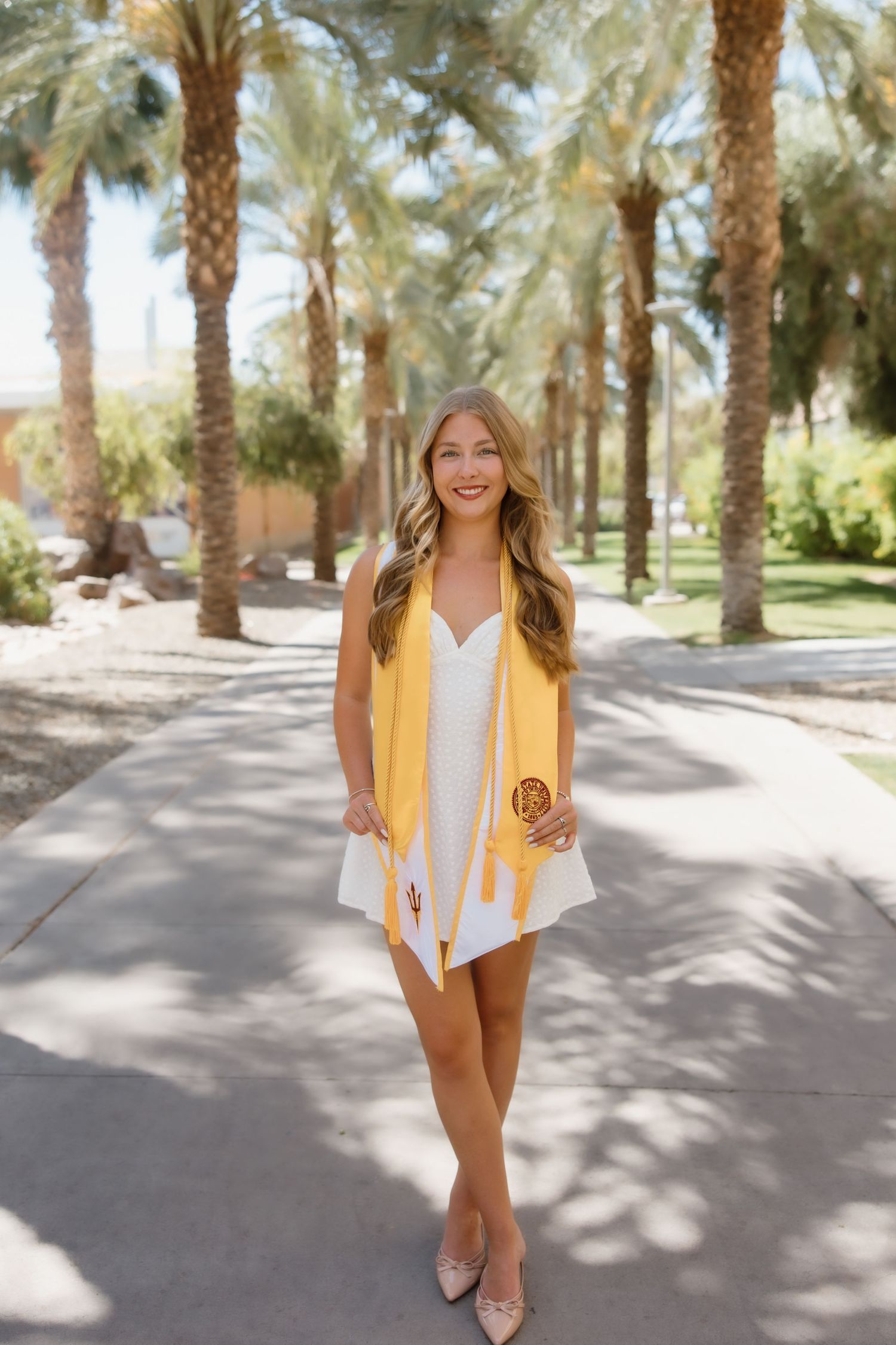 A smiling graduate in a white dress and gold sash poses on a palm tree-lined path outdoors.