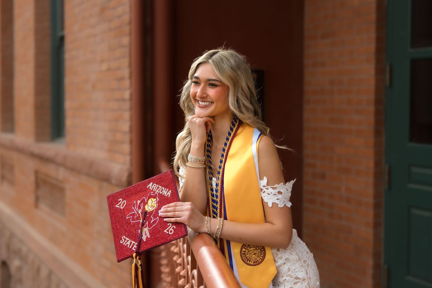 Smiling blonde college graduate holding Arizona State University cap and wearing gold sash outdoors near brick building.