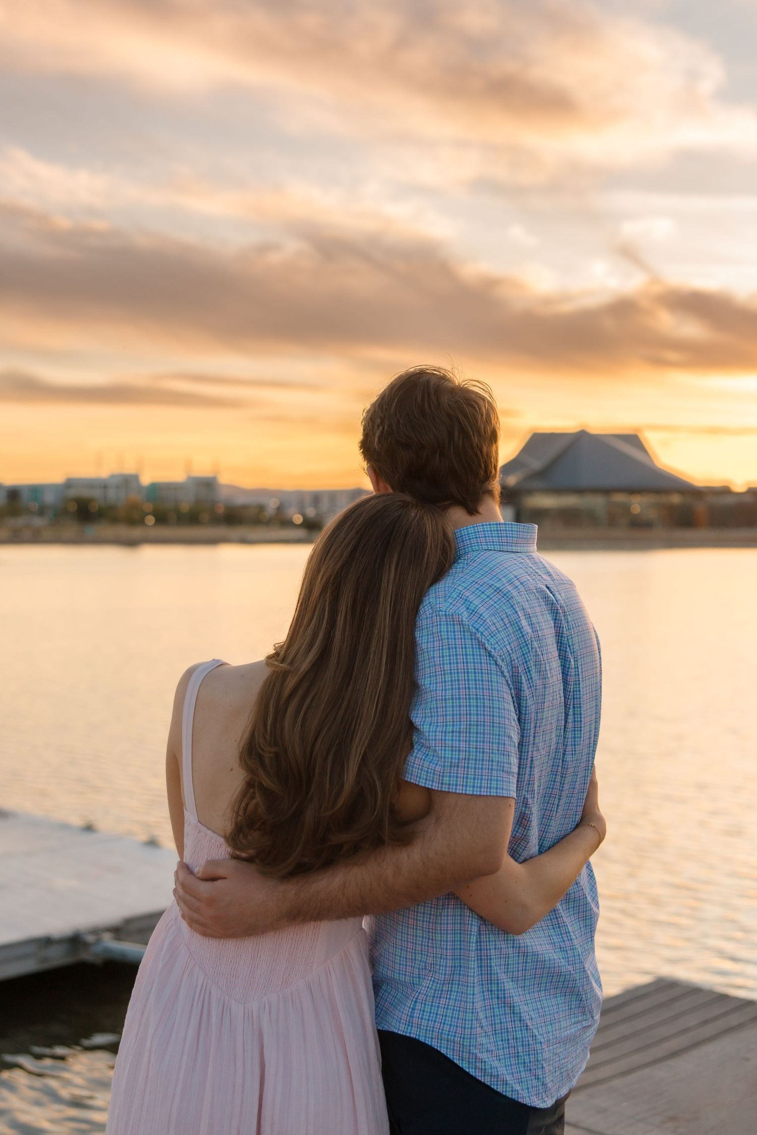 Couple standing arm in arm on a dock, gazing at a vibrant orange sunset over the water.