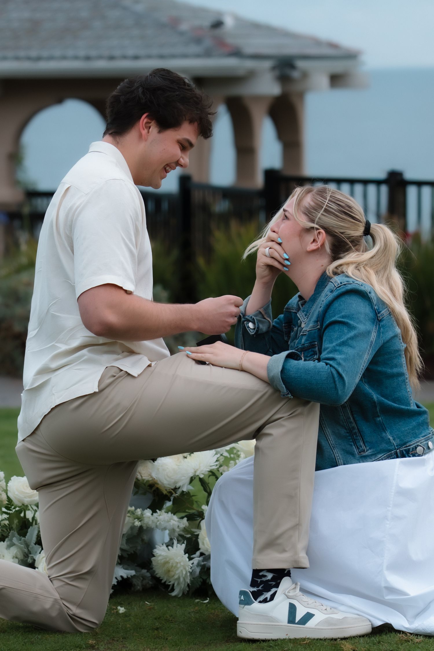 Man on one knee proposing to woman outdoors near ocean with white floral arrangement.