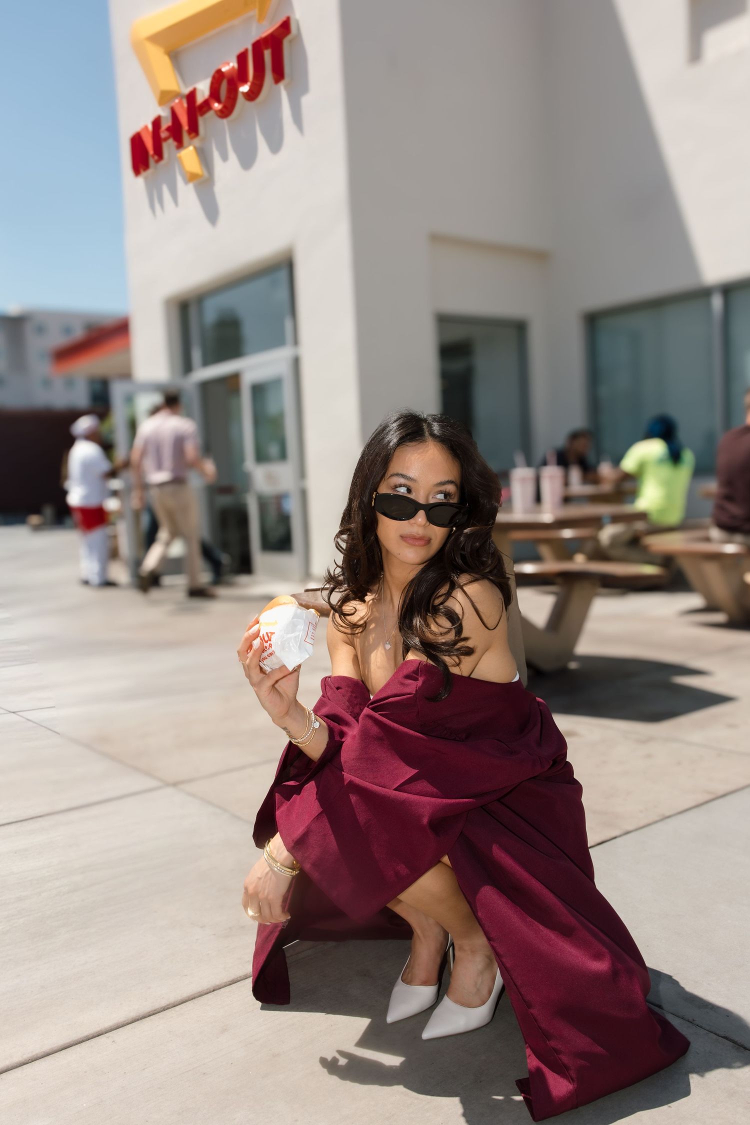 Female graduate in burgundy robe and sunglasses crouches holding an In-N-Out burger outside the restaurant.