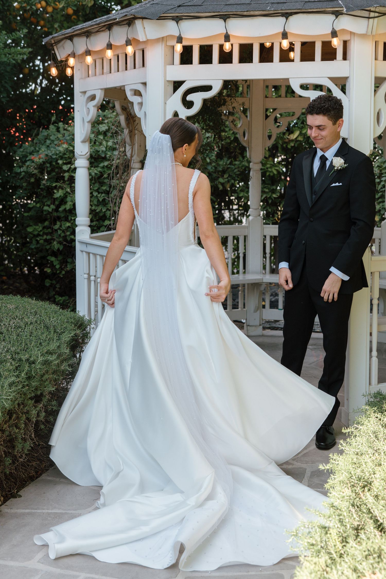 Groom in black tuxedo smiles as bride in flowing white gown with veil turns at garden gazebo.