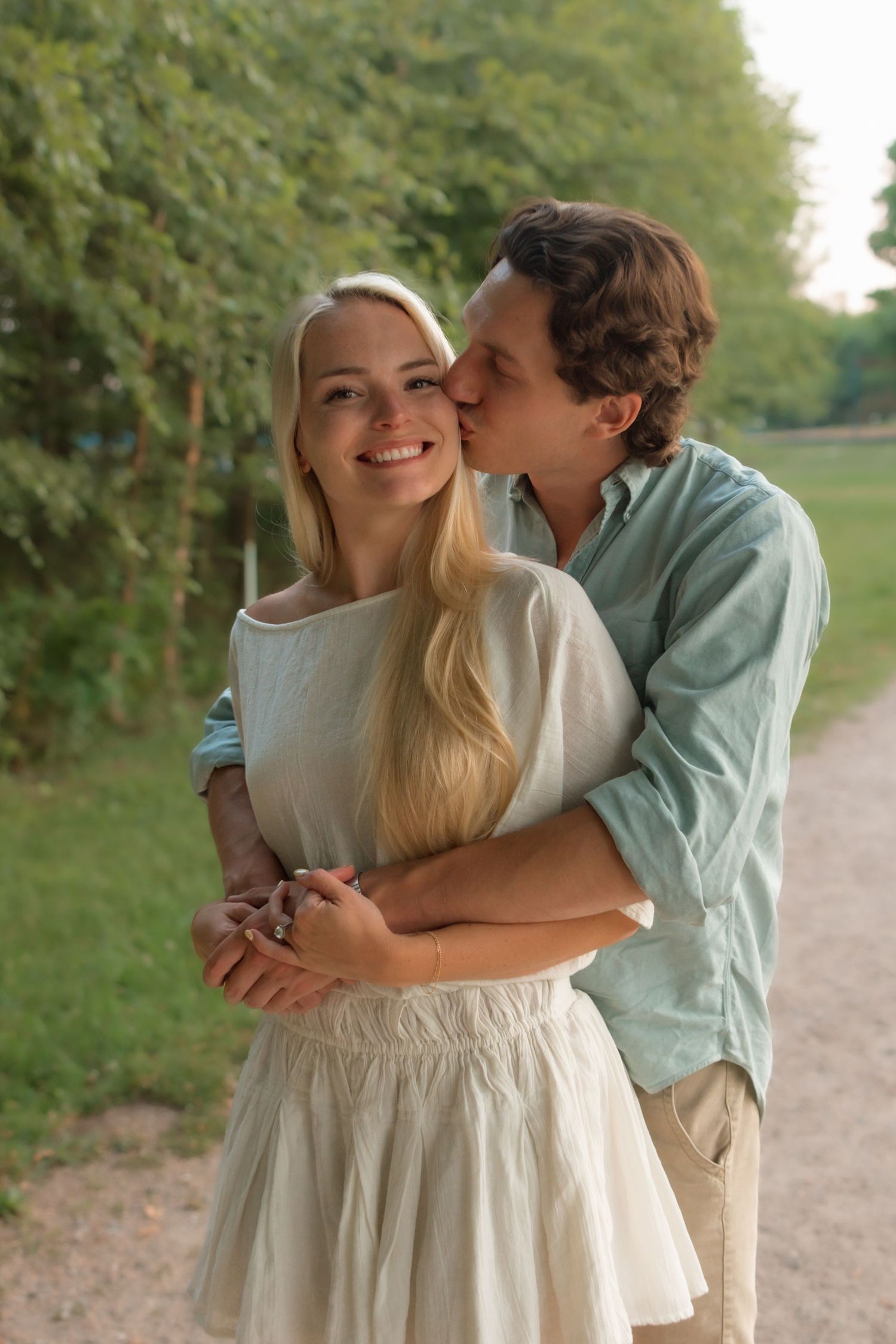 A man kisses a smiling woman on the cheek while embracing her on a park path.