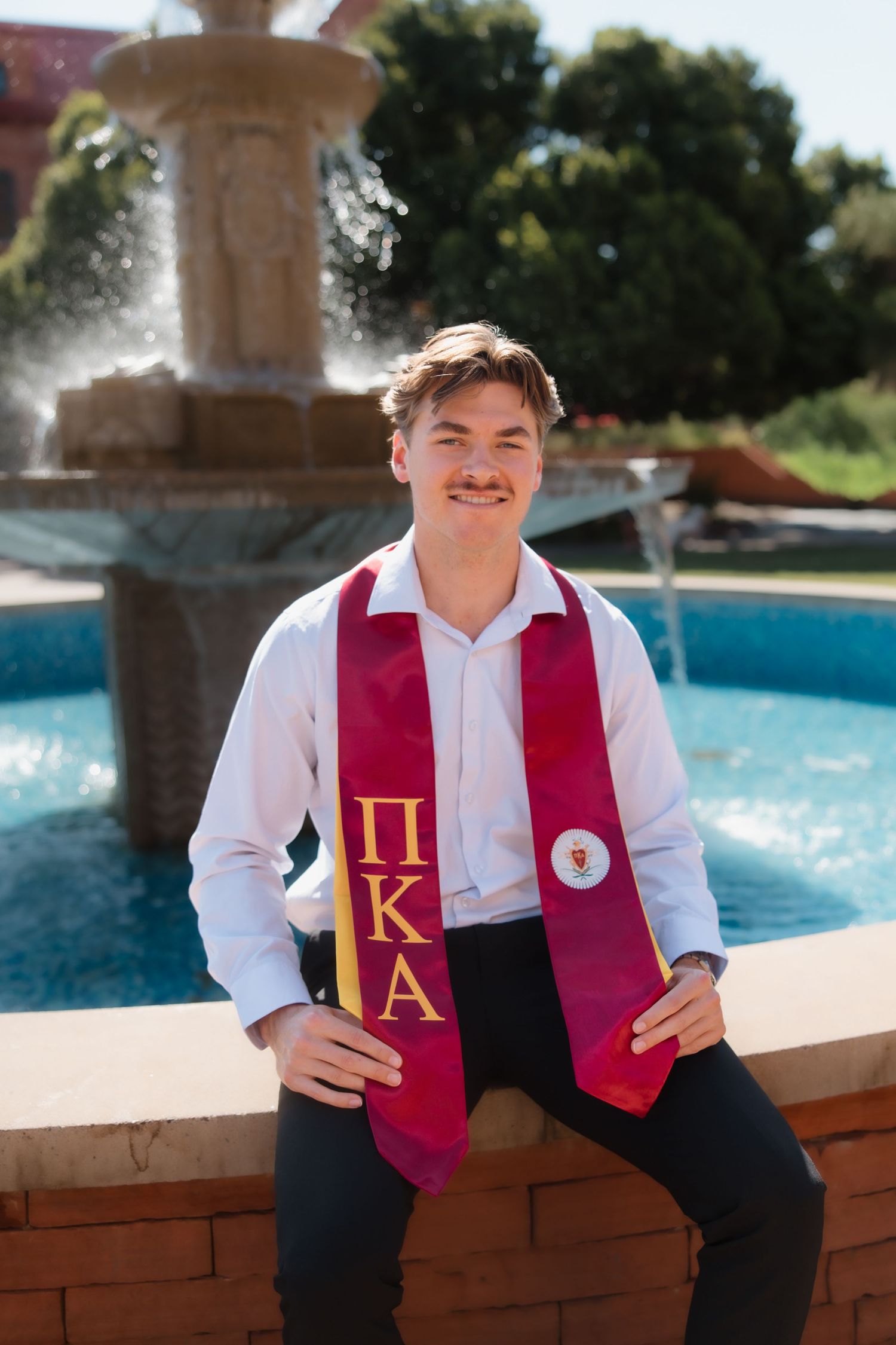 Young man in white shirt wearing Pi Kappa Alpha graduation stole seated by fountain for college graduation photos.