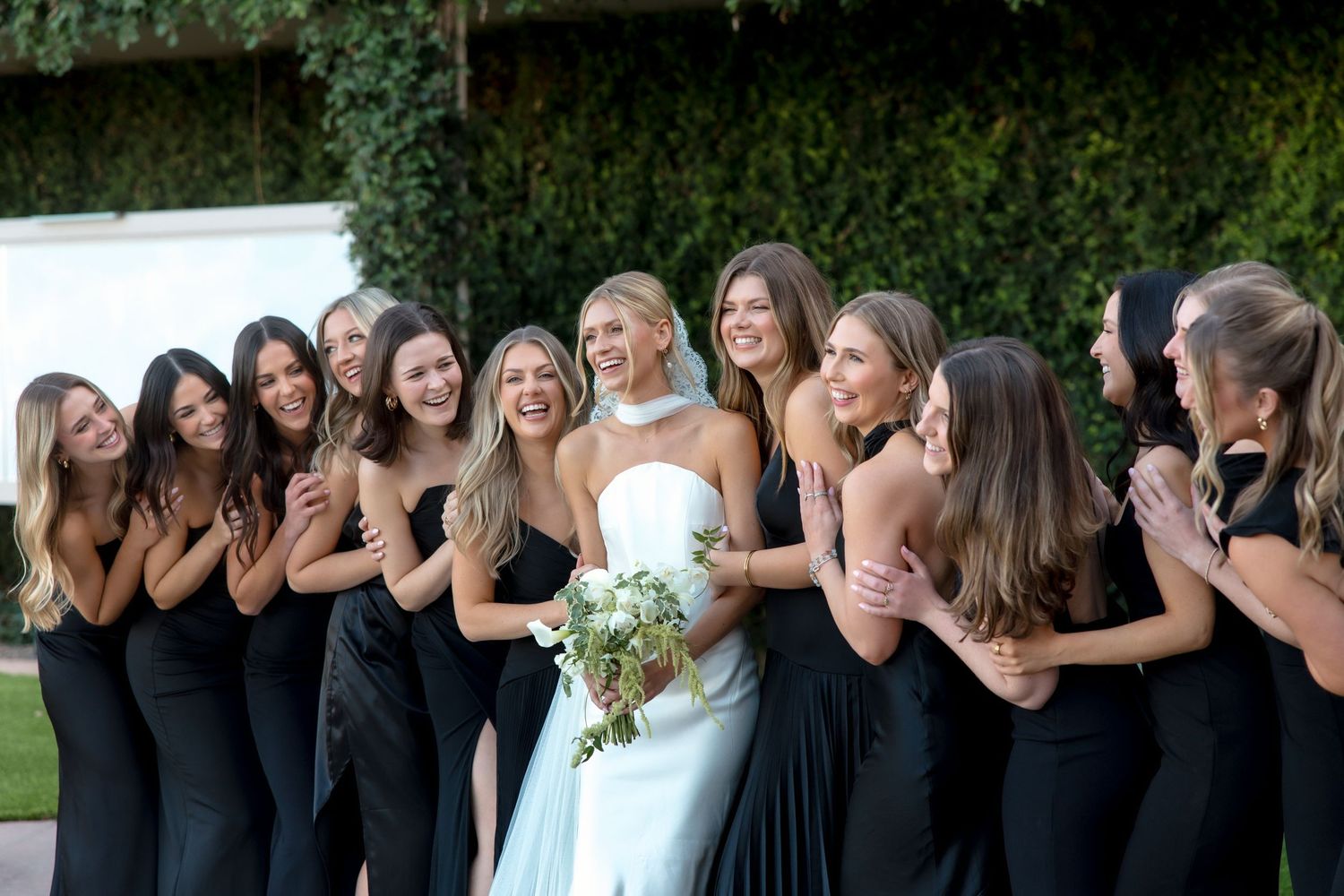 Bride in white halter gown laughs with bridesmaids in black dresses while holding white bouquet.
