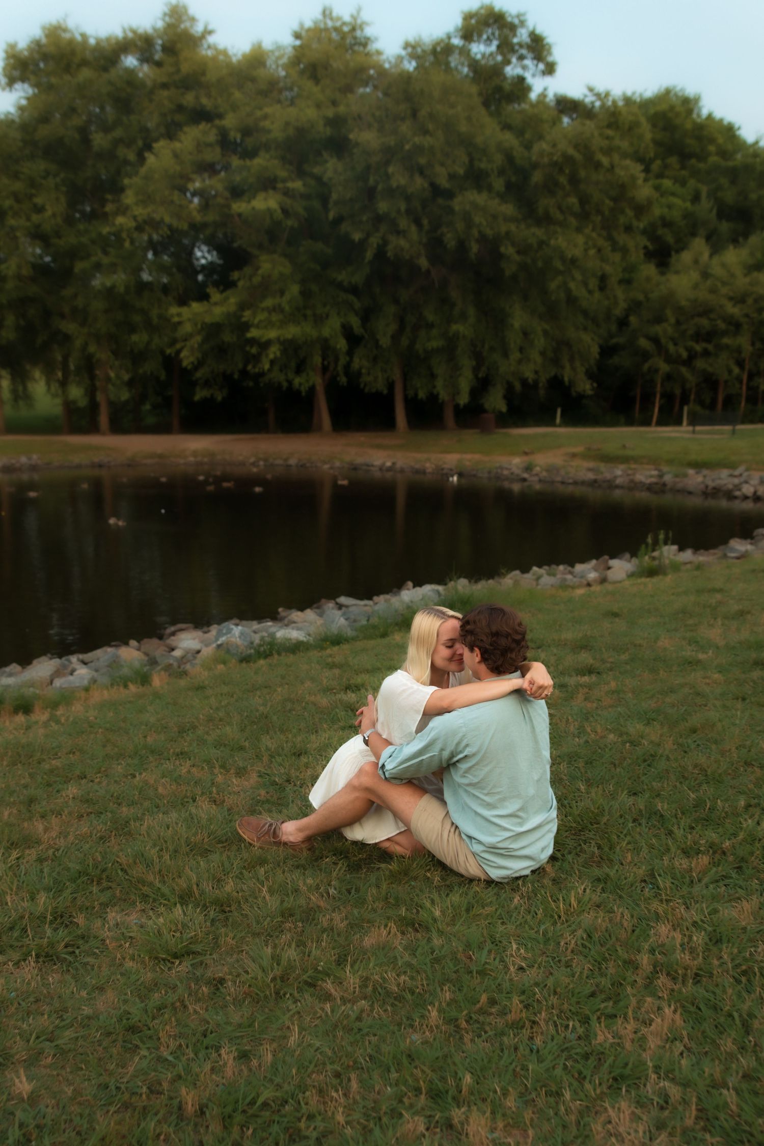 Loving couple embracing while seated on green grass beside a tranquil park lake.
