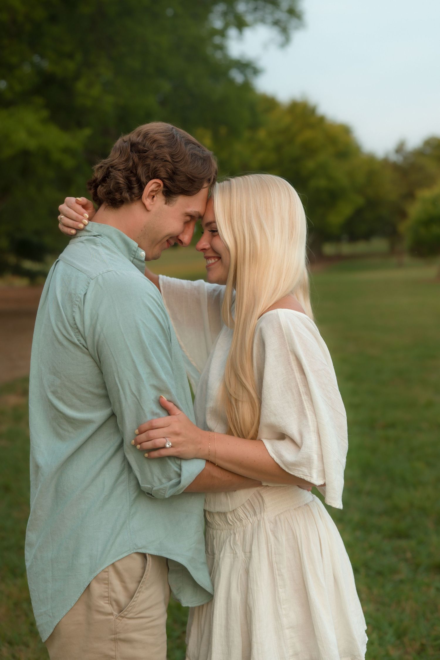 Romantic couple touching foreheads while embracing in a green park at dusk.
