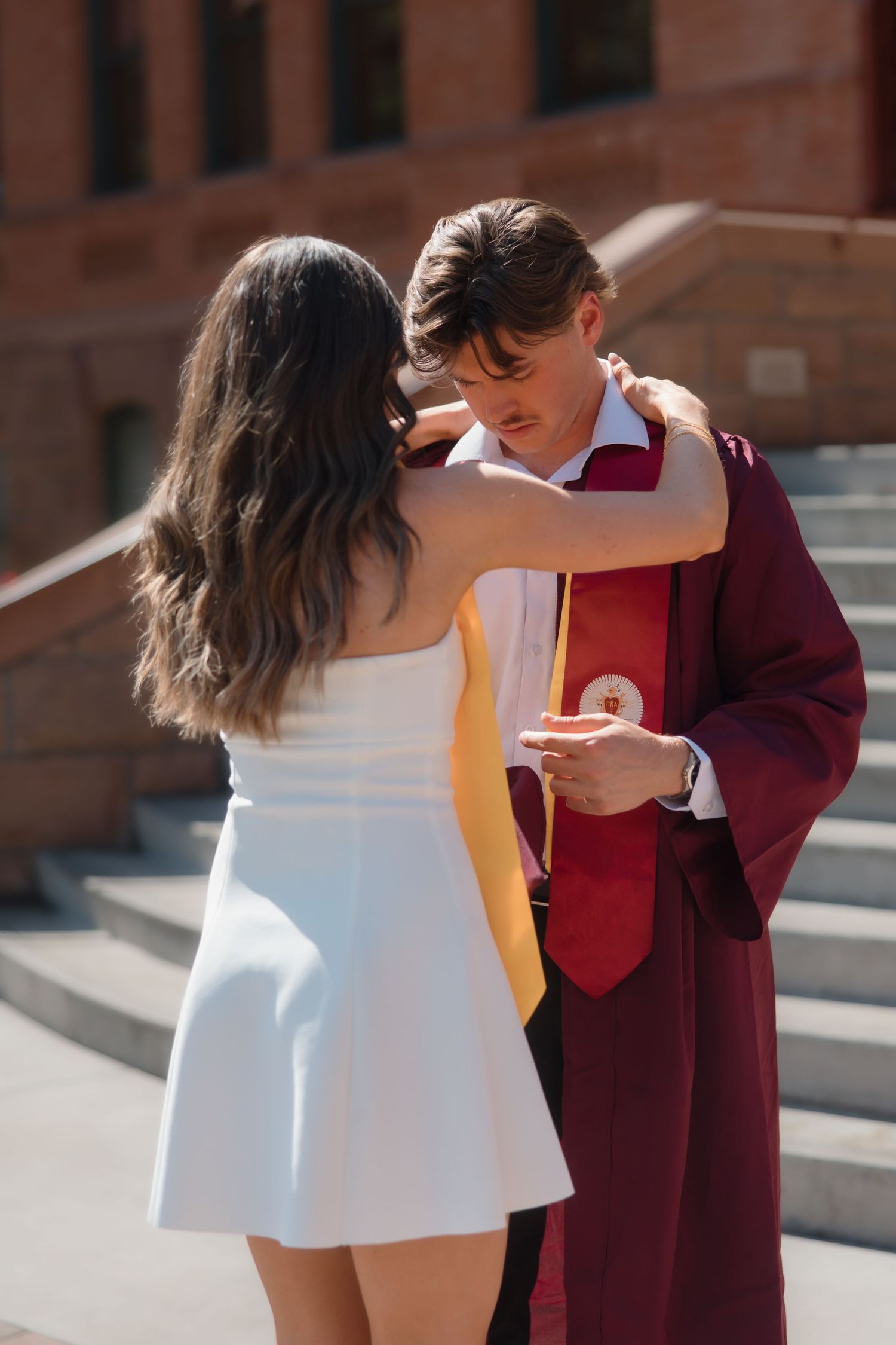 Graduate in maroon robe shares a tender moment with woman in white dress on sunny college campus steps.