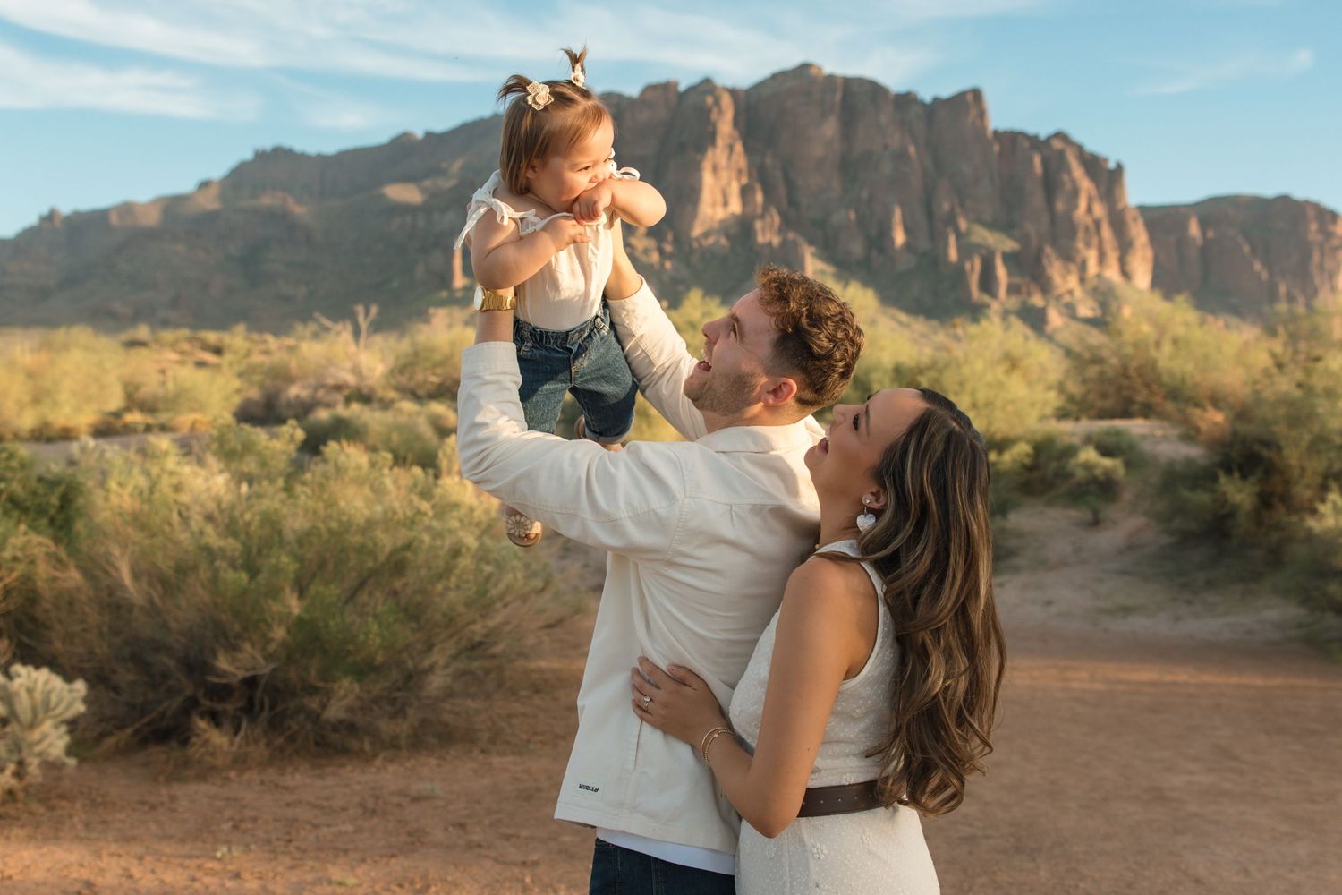 Father lifting joyful baby girl in the air while mother watches in a desert landscape.