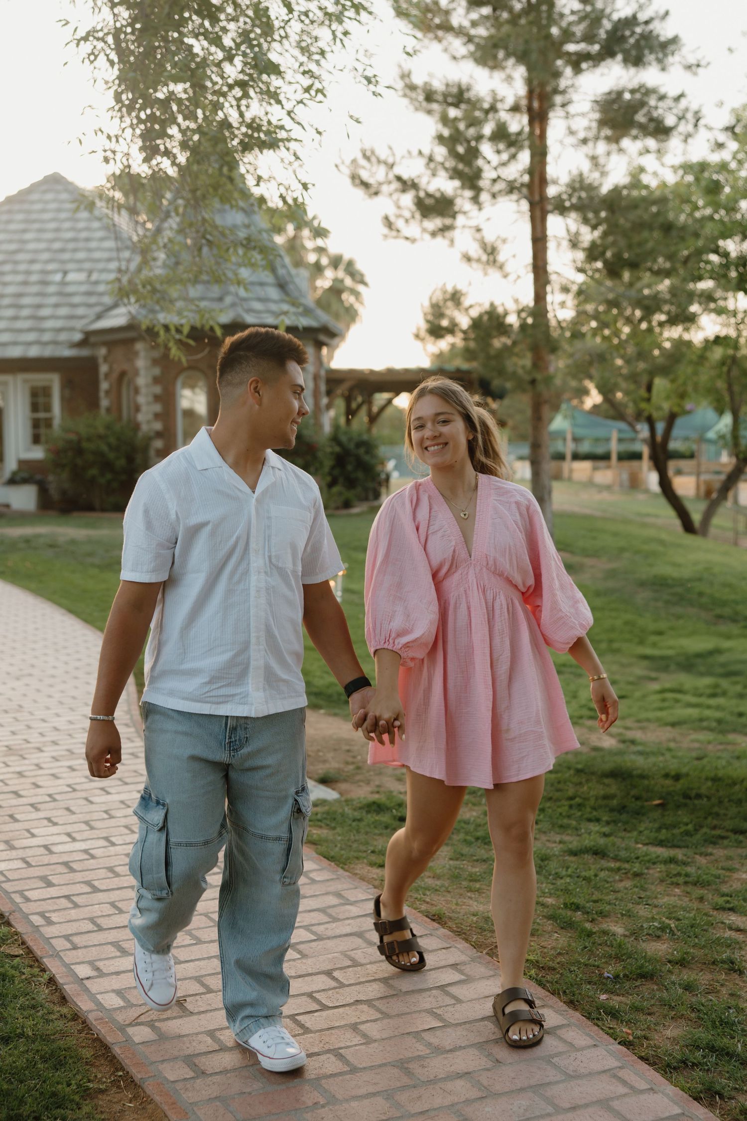 Couple holding hands walking on brick path in park, man in white shirt and jeans, woman in pink dress, smiling.