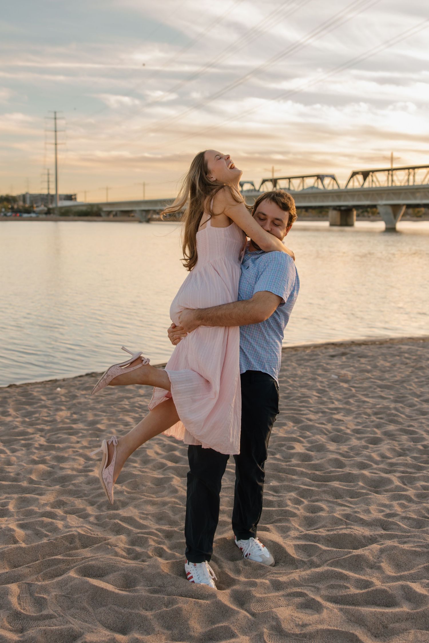 Couple embracing on sandy beach at sunset, man lifting woman in pink dress near river with bridge backdrop.