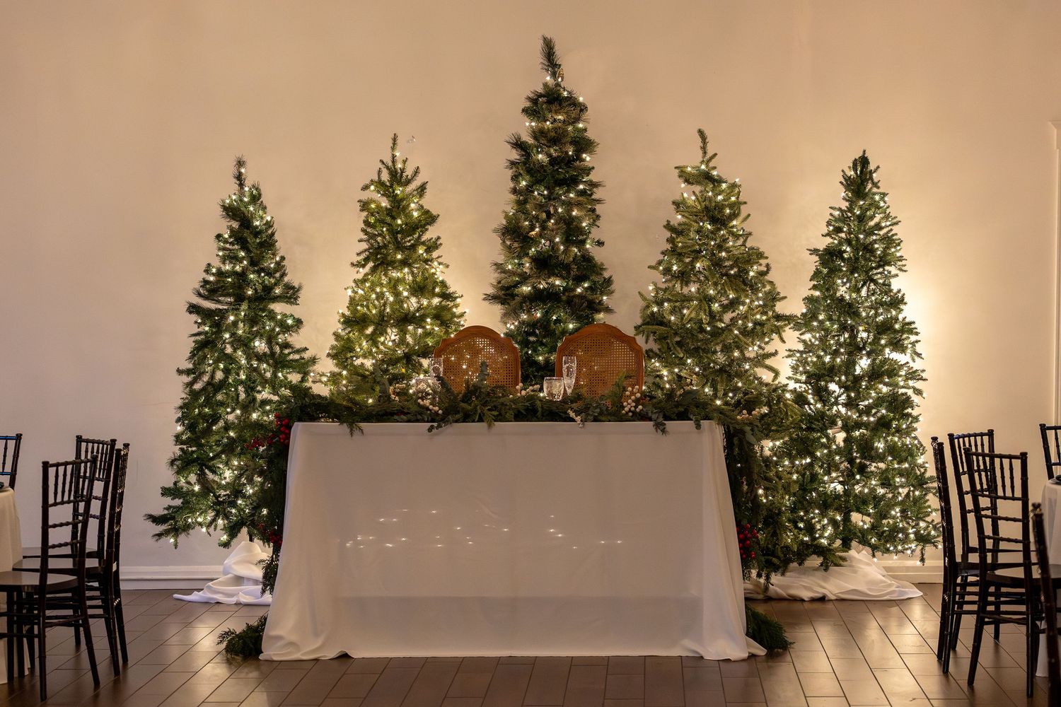 Festive wedding ceremony space with illuminated Christmas trees and white draped sweetheart table.