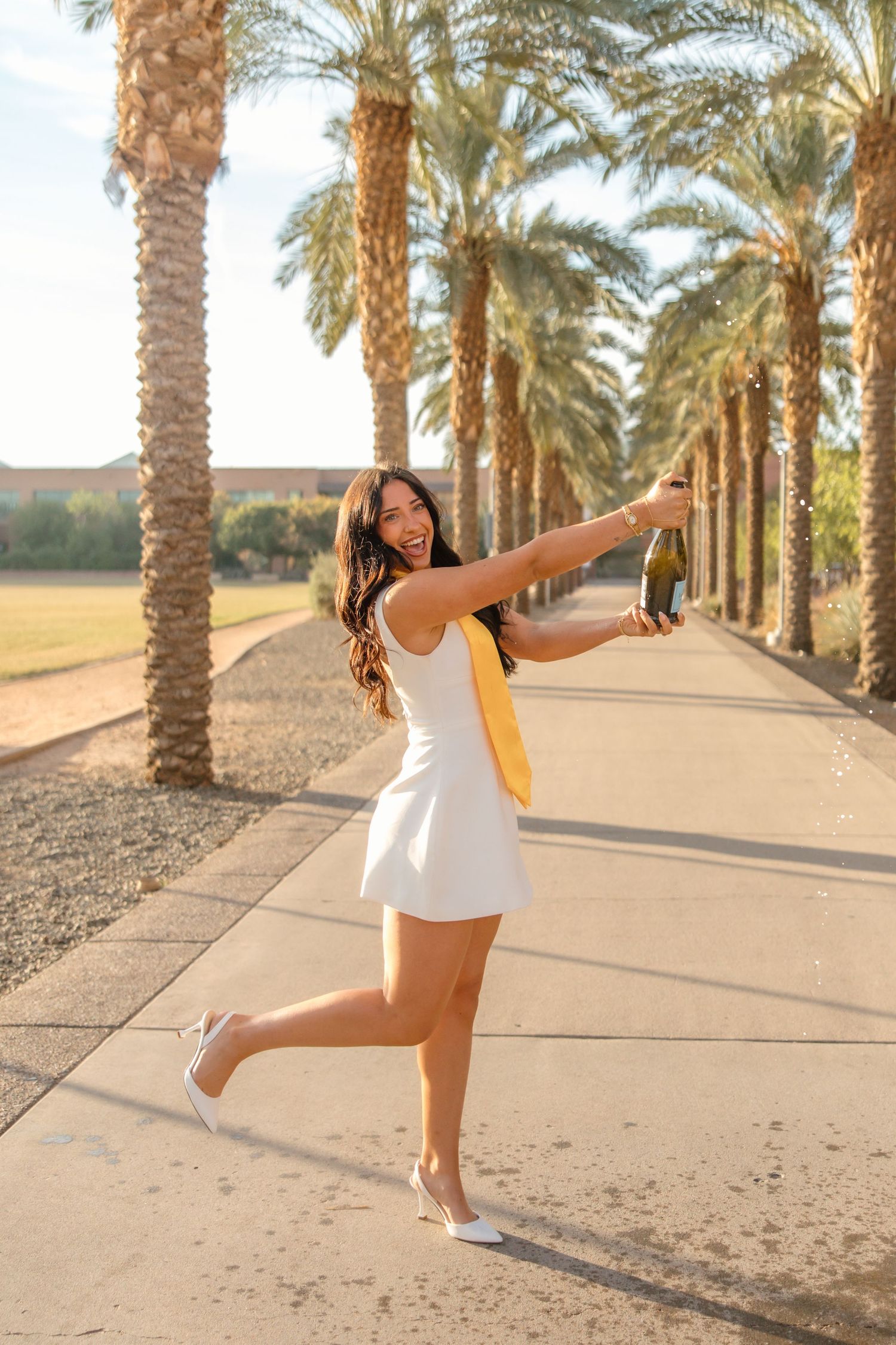 Young woman in white dress holding champagne bottle celebrates outdoors along palm tree-lined pathway in heels.