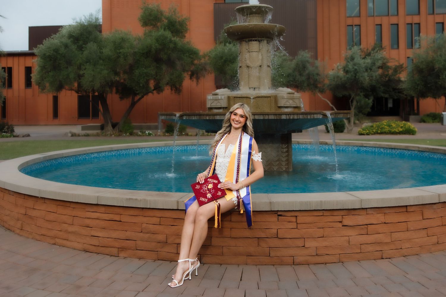 A smiling female graduate in white gown with colorful sashes poses by a fountain on a sunny campus.
