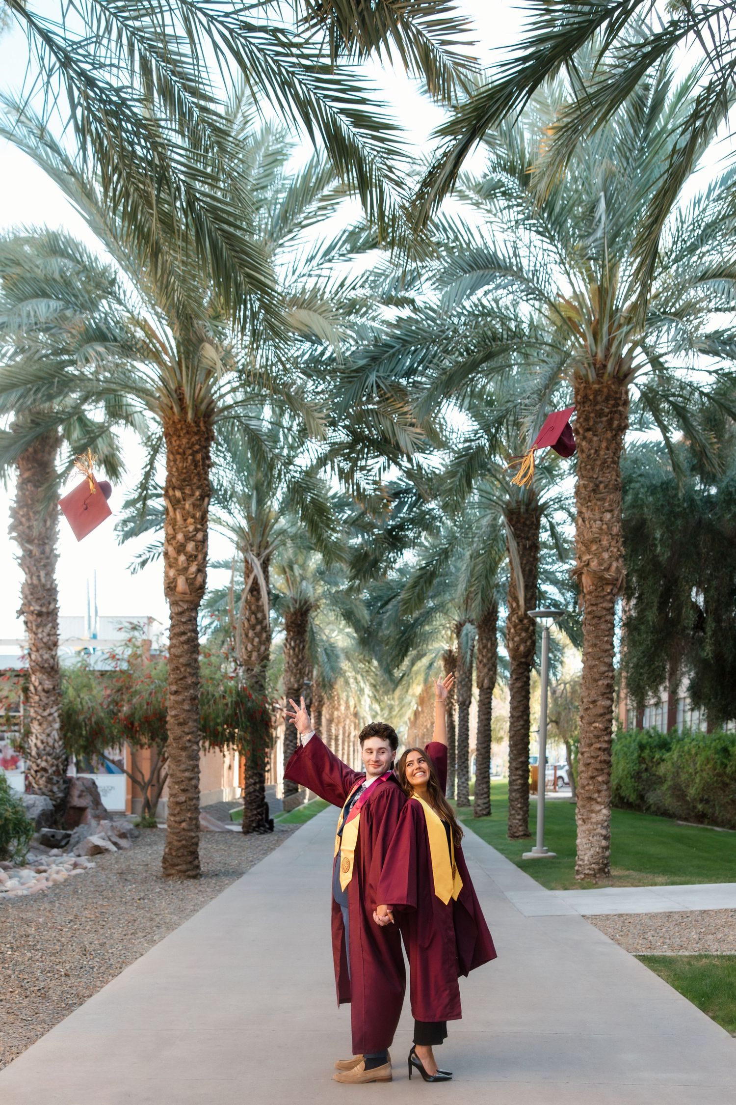 Two graduates in maroon caps and gowns celebrate outdoors along a palm tree-lined walkway.