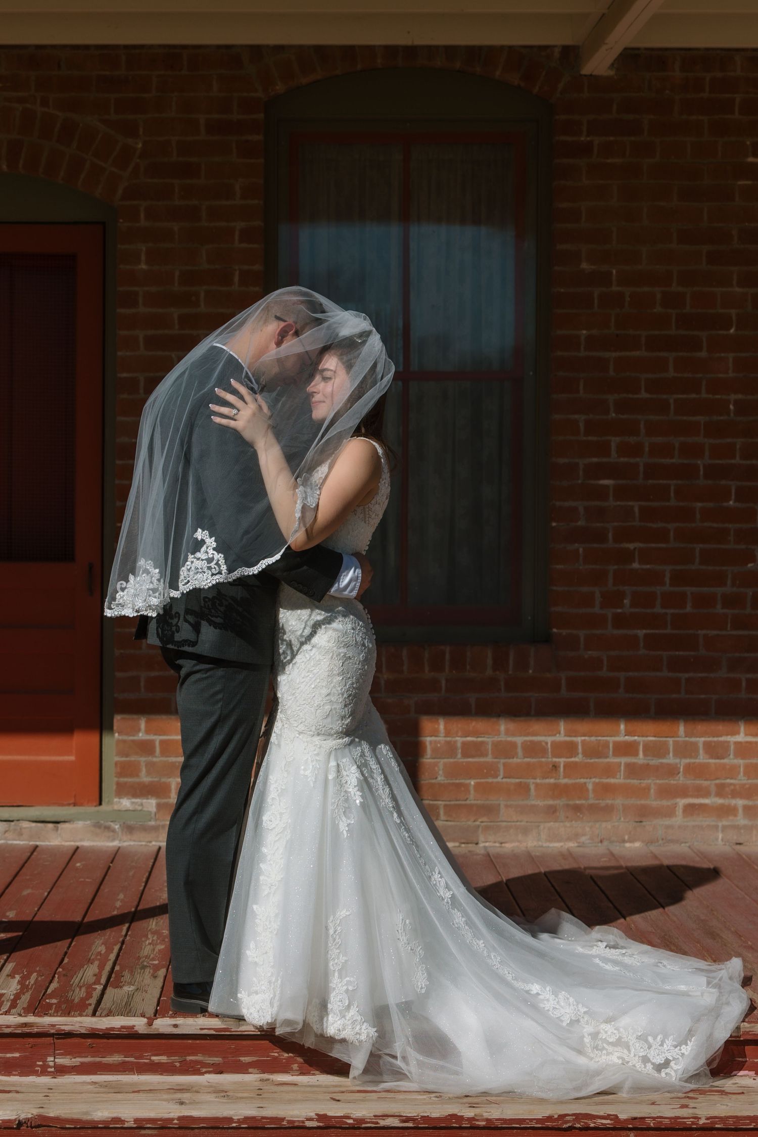 A bride and groom share a romantic kiss under a lace veil outside a brick building.