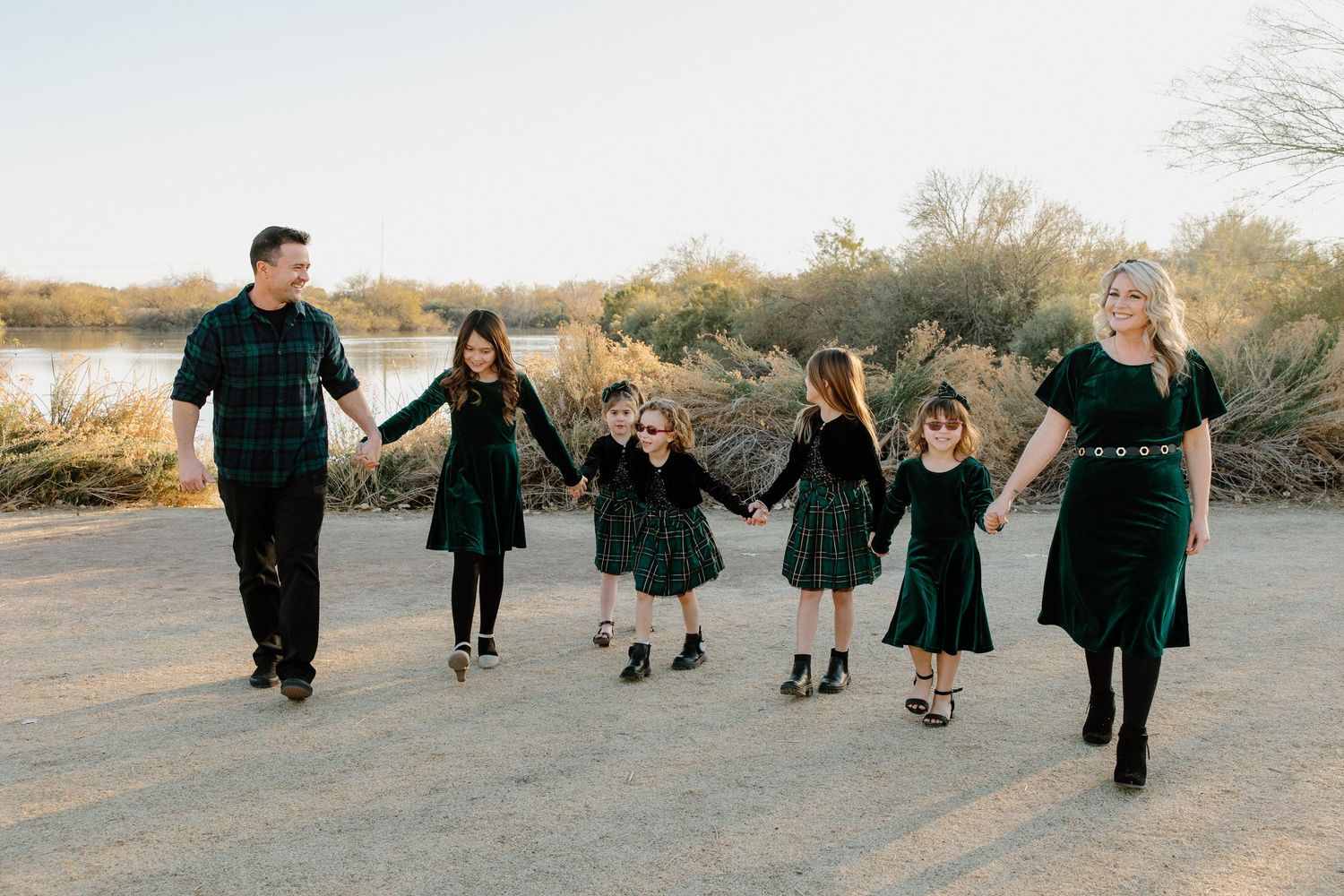 Family of six in matching green plaid outfits walking hand-in-hand outdoors near a lake during golden hour.