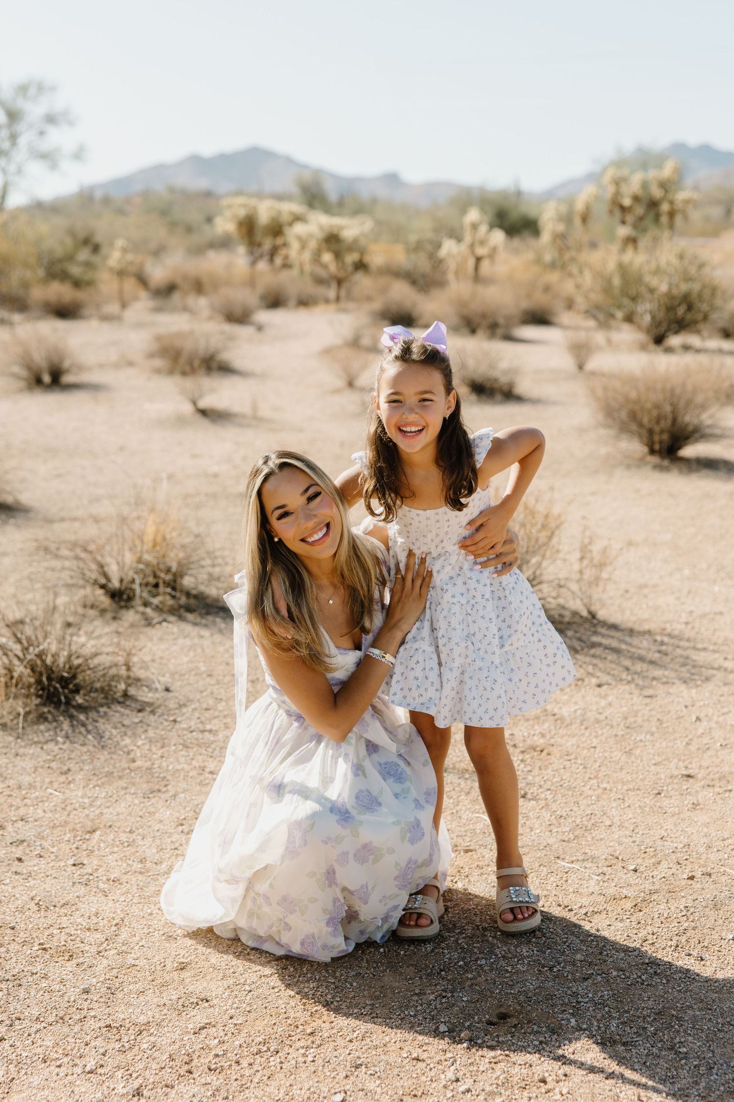 A woman and young girl in floral dresses pose together in a desert landscape with cacti and mountains.