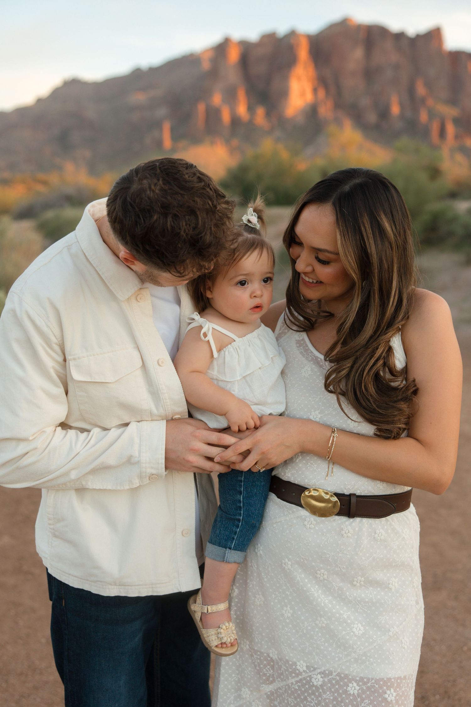 Family portrait with parents holding toddler girl in white dress, desert mountains at golden hour.