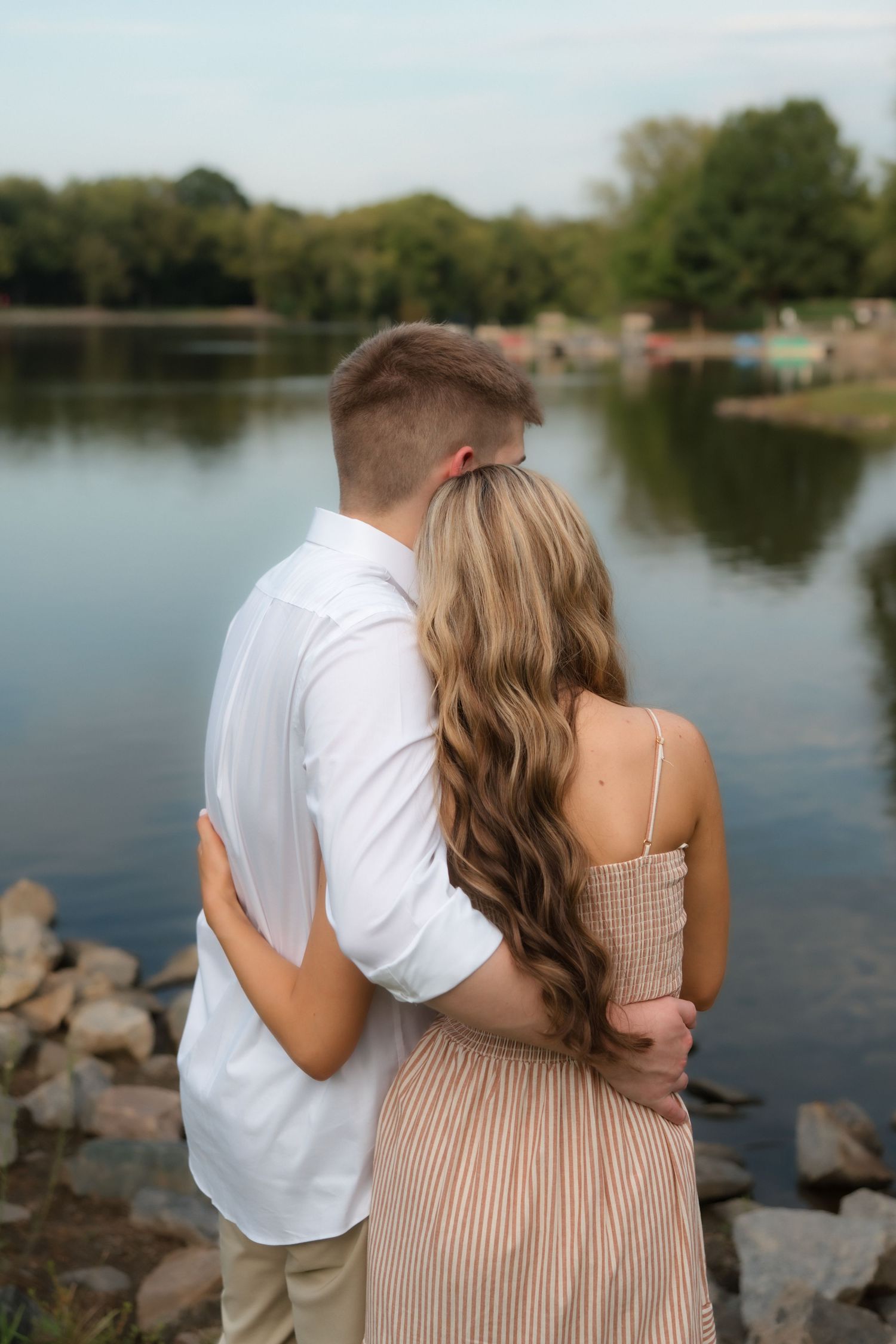 Couple embracing by a serene lake, woman in striped dress with long wavy hair, man in white shirt, rocky shore.