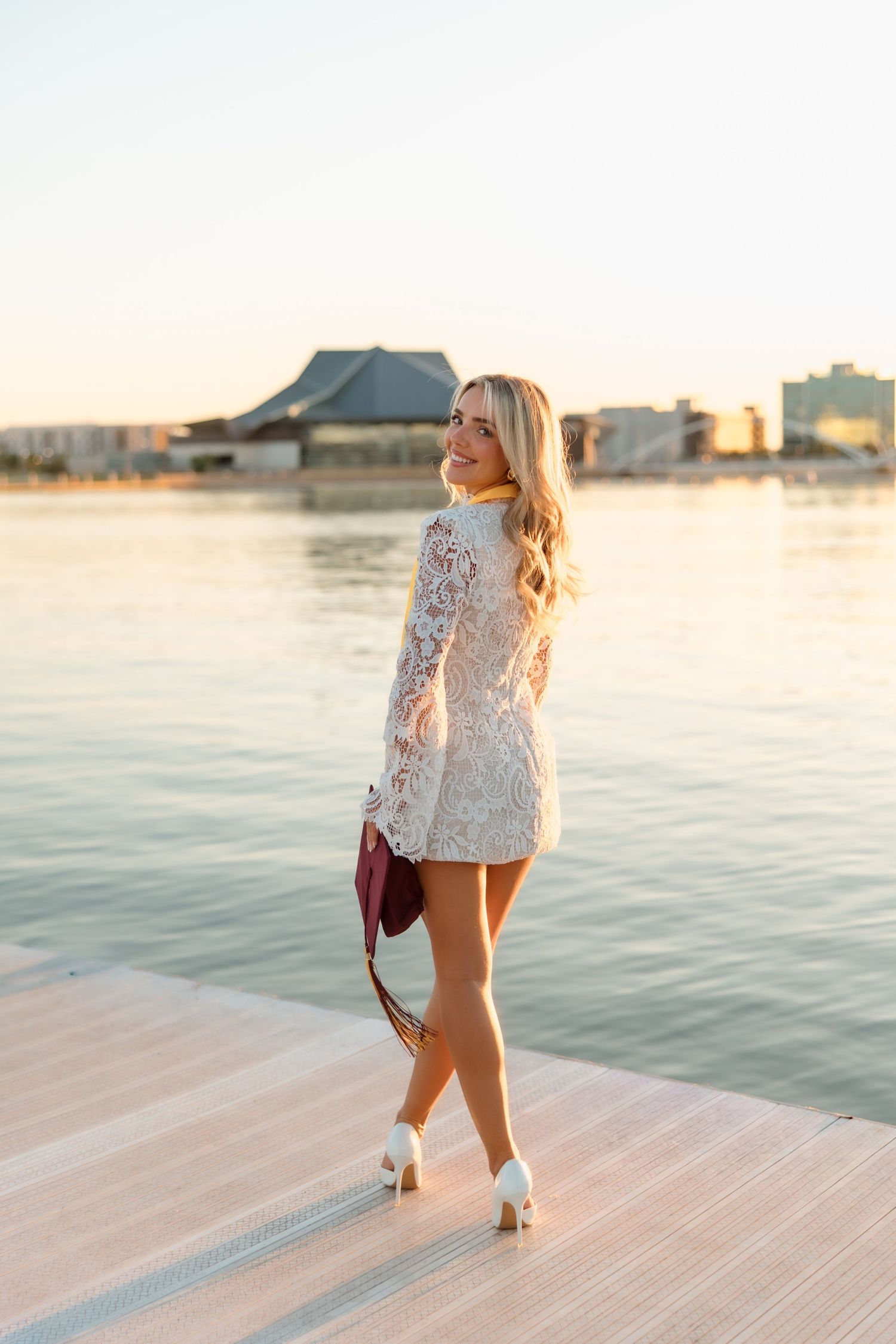 Blonde woman in white lace mini dress and heels posing on waterfront dock at golden hour sunset.