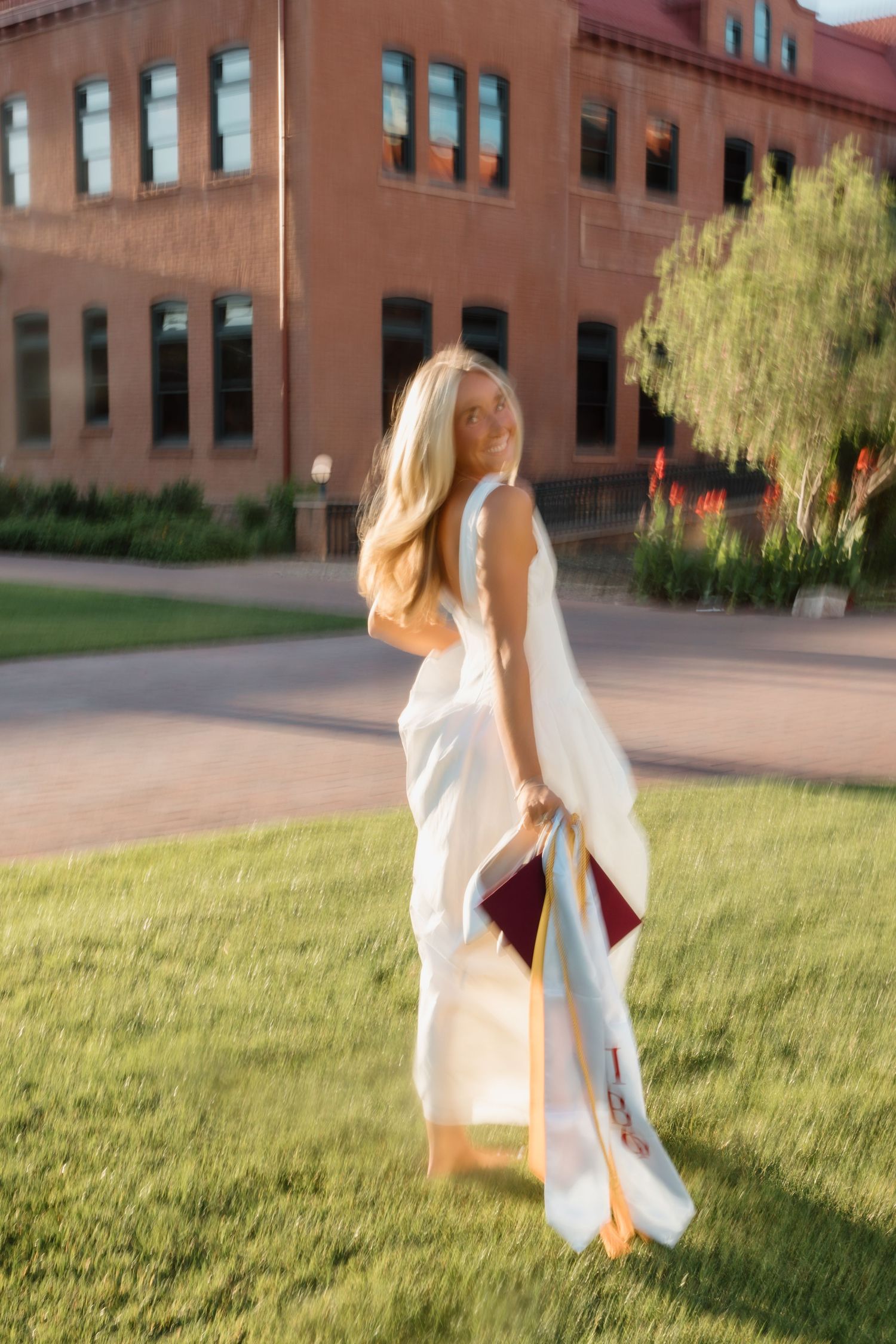Blonde woman in white dress holding graduation cap on green campus lawn with brick building background.