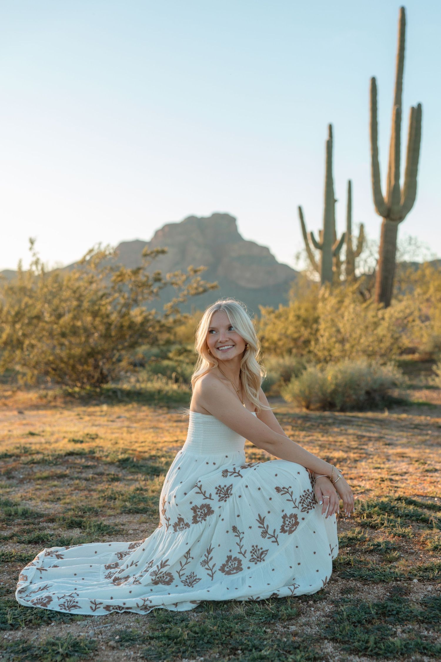 Smiling blonde woman in floral white dress sitting in Arizona desert with saguaro cacti and mountain at golden hour.