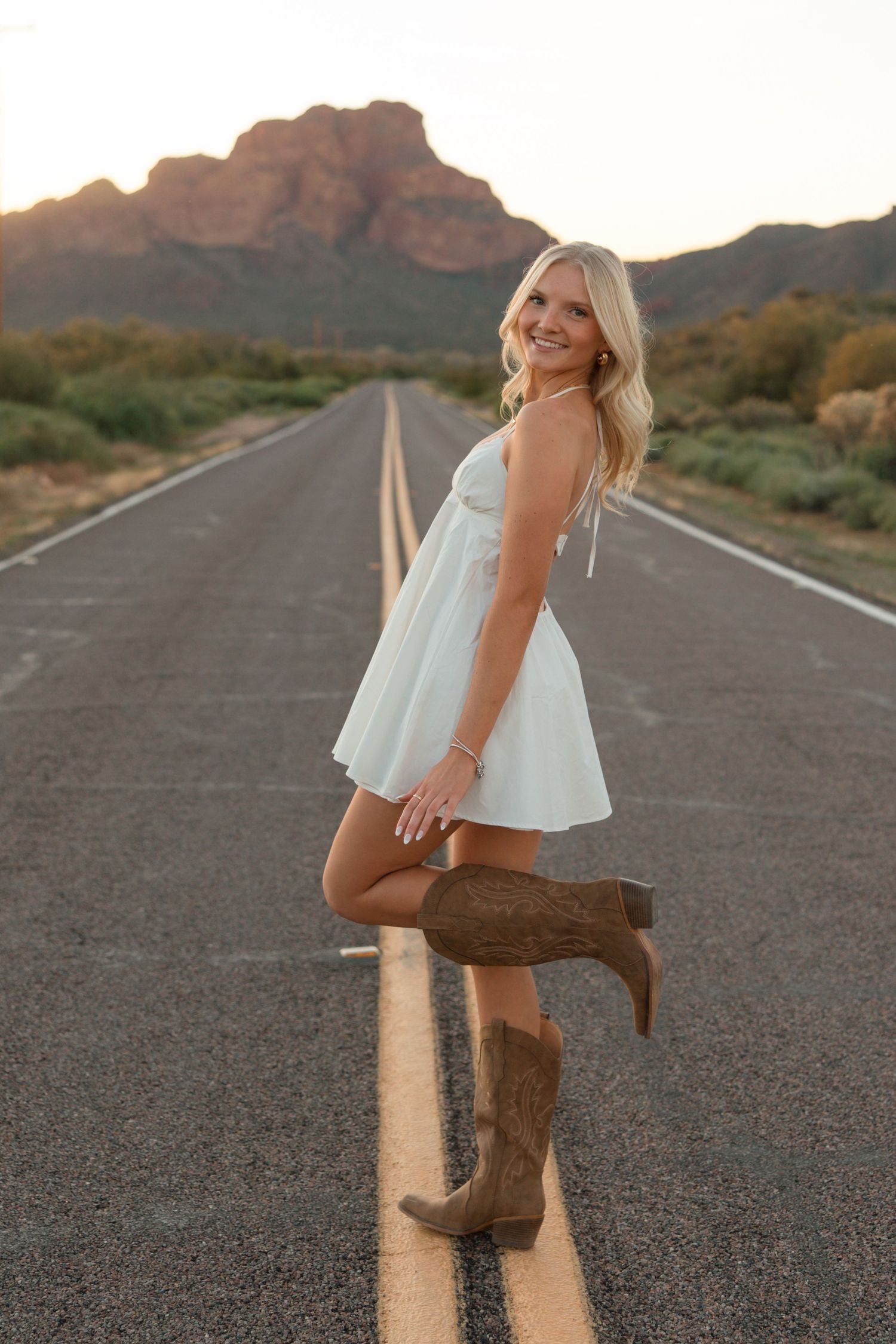 Blonde woman in white dress and cowboy boots posing on desert road with mountain backdrop at sunset.