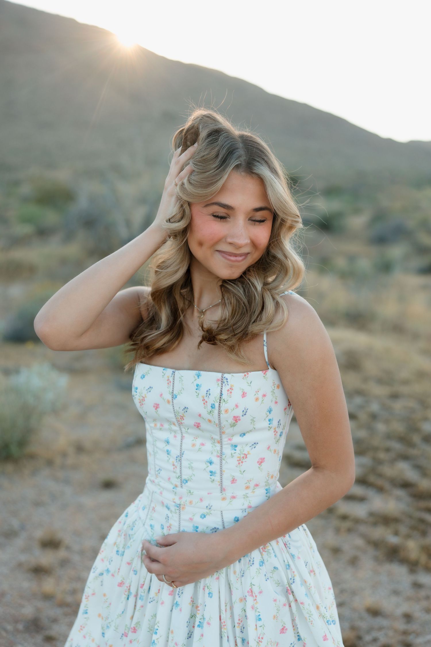 Young woman in floral sundress poses outdoors in desert landscape with golden sunset light behind mountains.