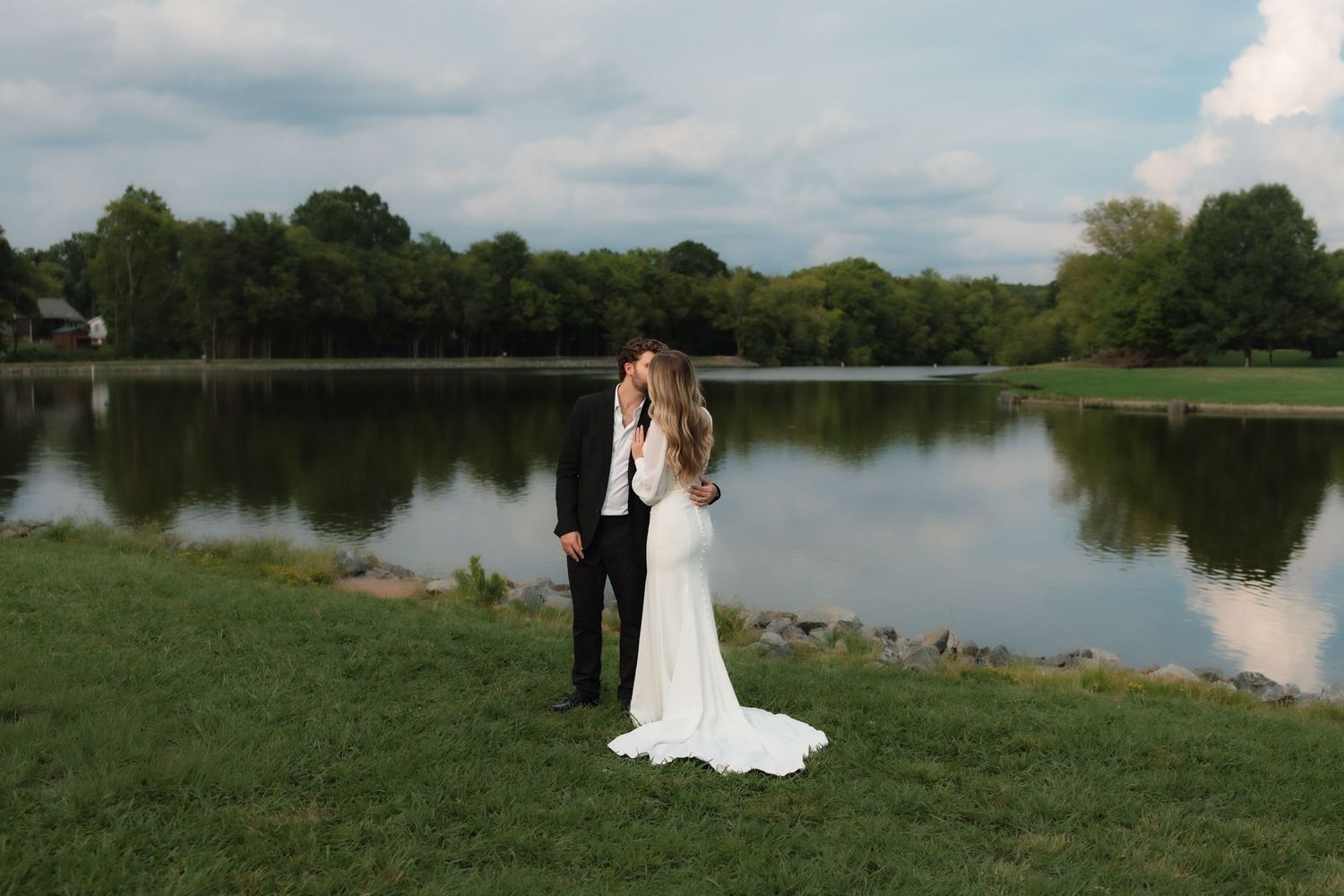 Bride and groom kissing beside a serene lake, bride in white gown with long train on lush green grass.