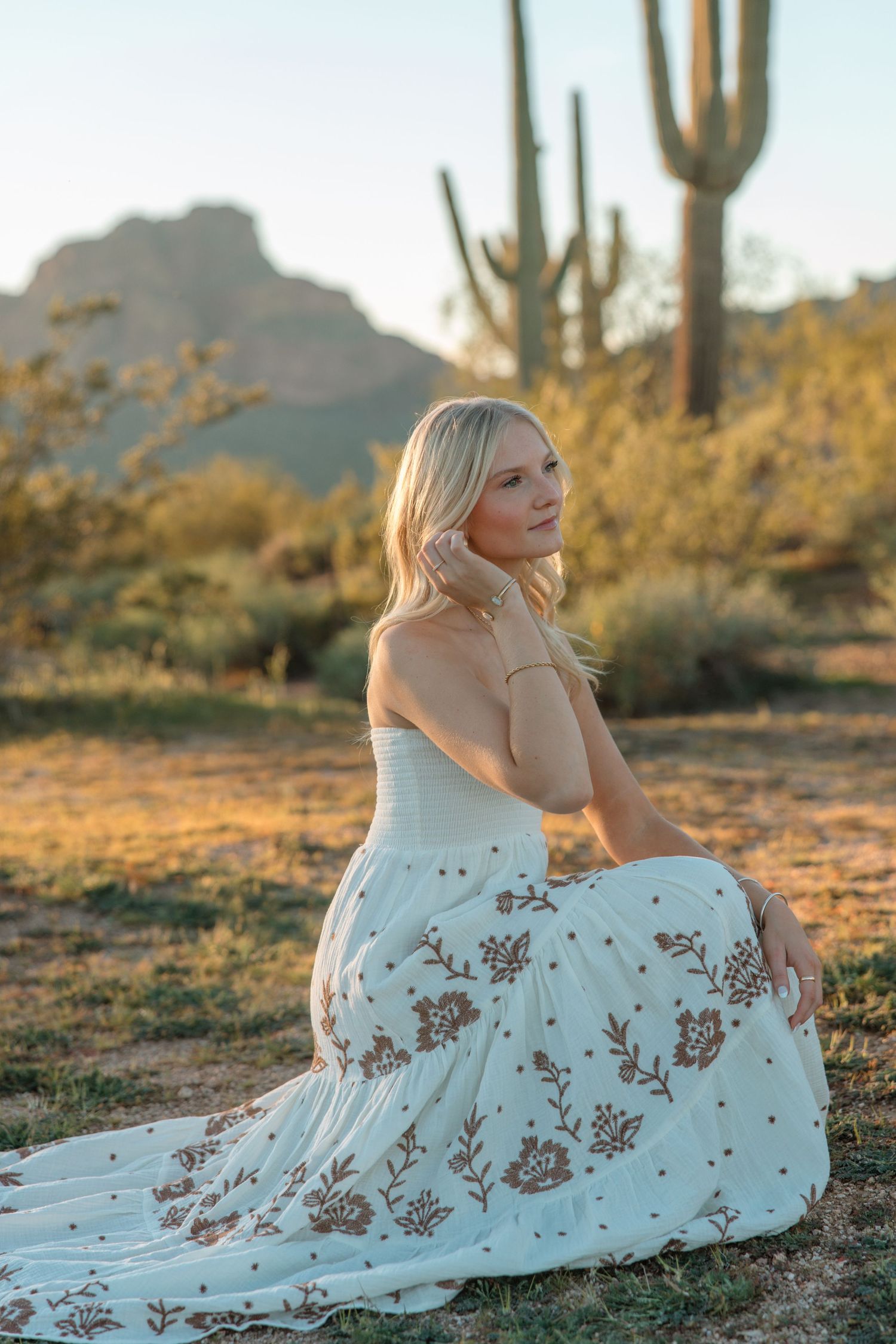 Blonde woman in white floral maxi dress sits in desert landscape with saguaro cactus at golden hour.
