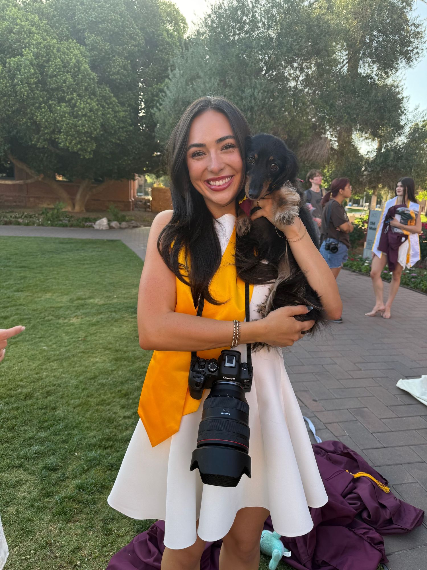 Smiling graduate in orange sash holds puppy and DSLR camera at outdoor university graduation ceremony.