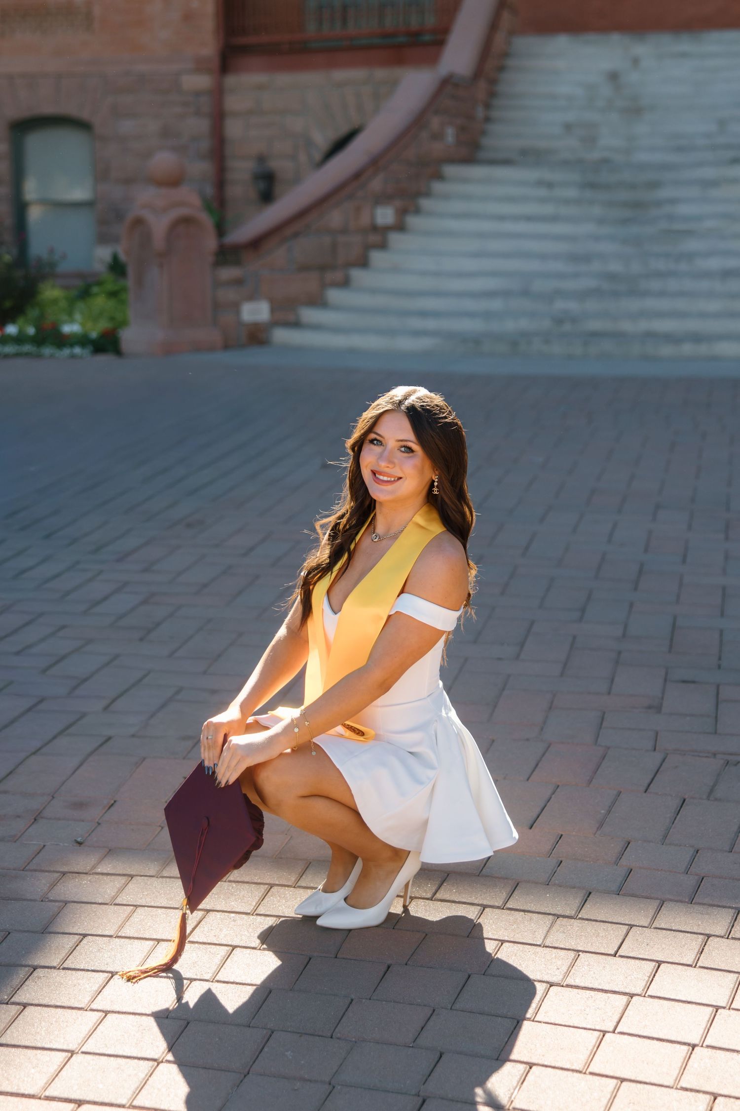 Smiling graduate in white dress and yellow stole crouching with maroon cap on brick plaza.