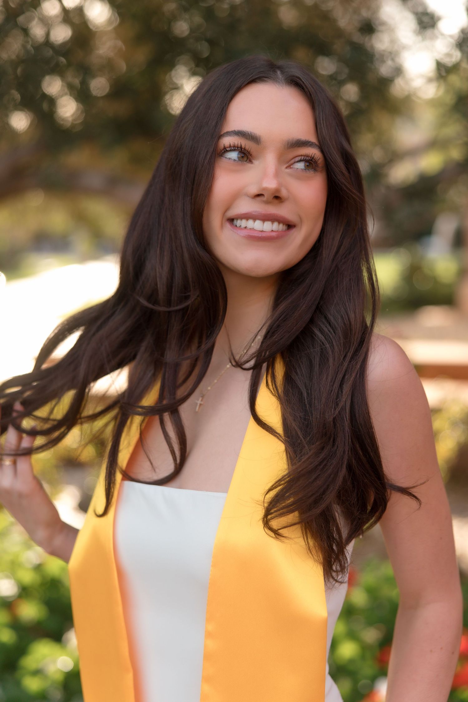 Smiling brunette female graduate wearing gold graduation stole in white dress with hair blowing outdoors.